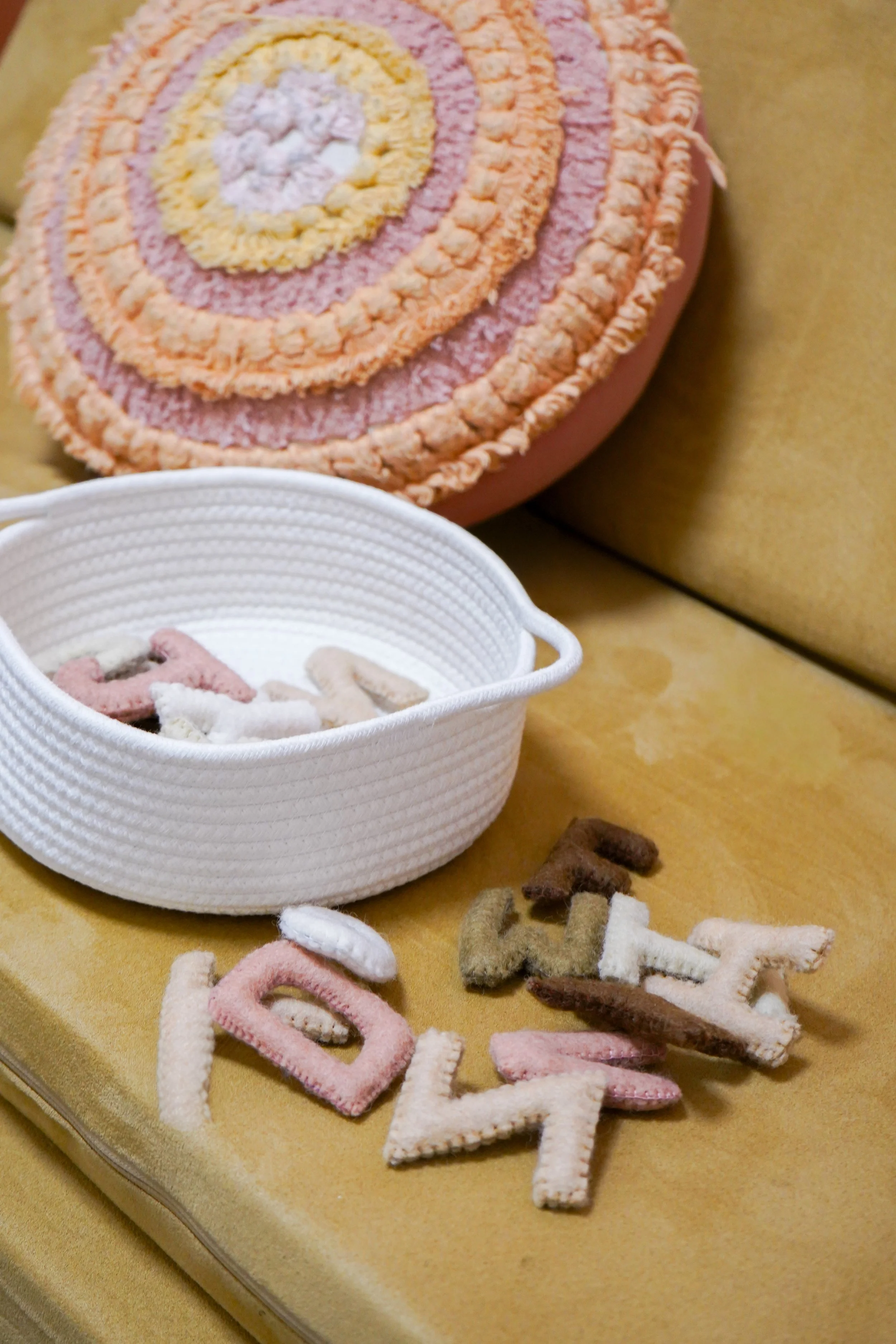A decorative cake with a pink, yellow, and white swirl design, a white basket containing felt alphabet letters, and several felt letter decorations scattered on a gold-colored surface.