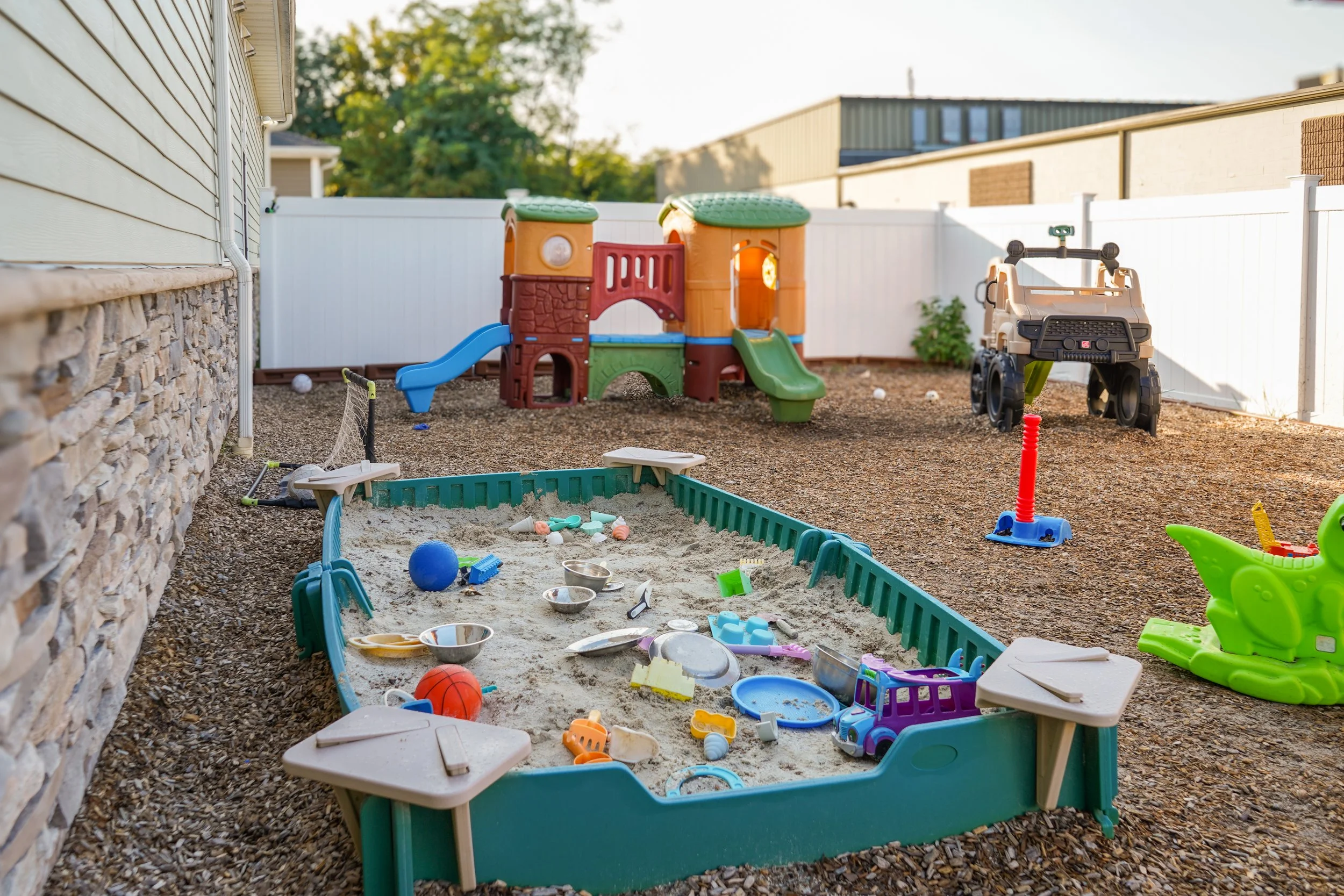 Backyard with kids' play area including a sandbox with toys, a plastic slide and climbing structure, toy car, small sports net, and a ride-on green dinosaur