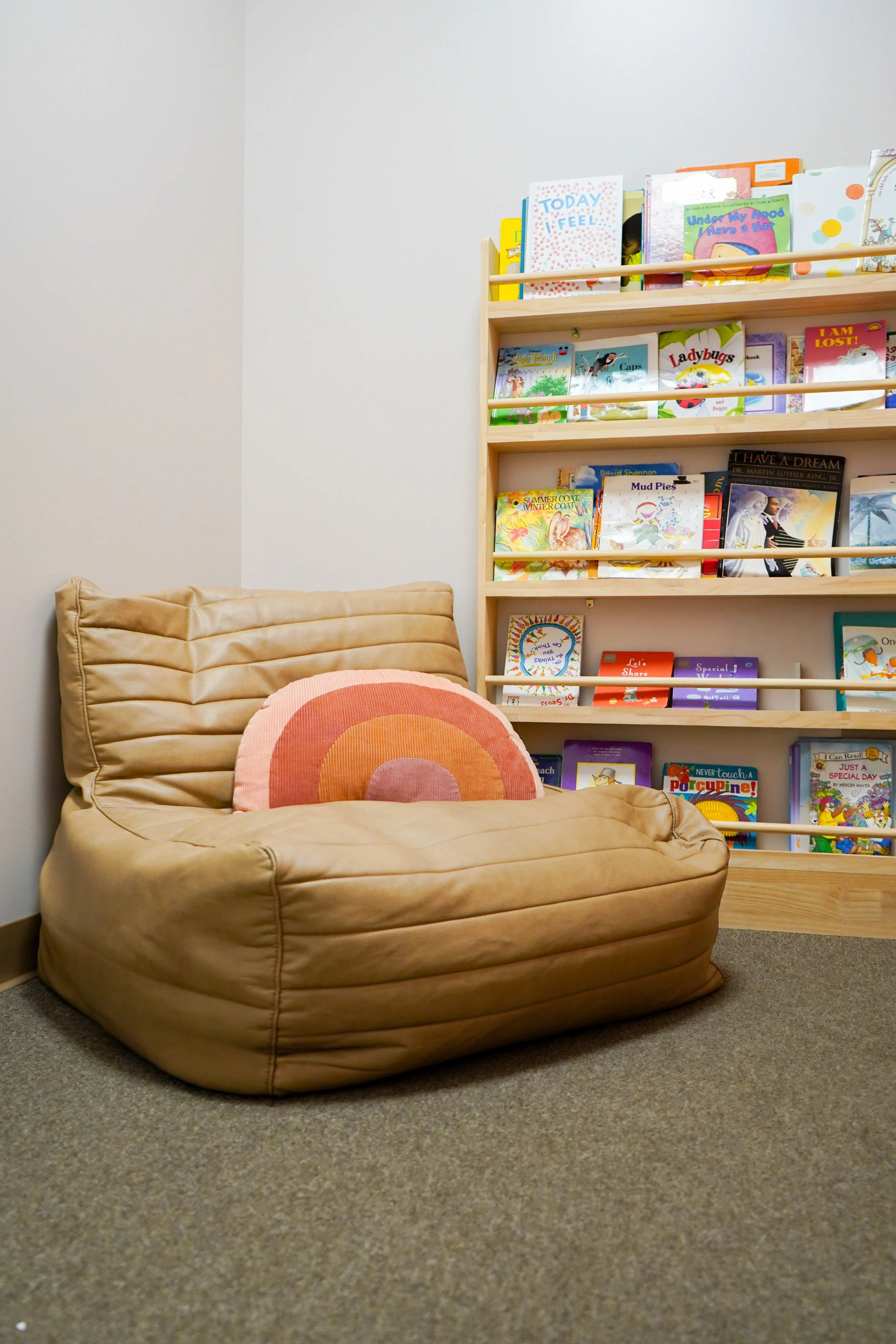 A beige, cushioned armchair with a pink and orange rainbow-shaped pillow on it, positioned next to a wooden bookshelf filled with colorful children's books in a cozy library or reading corner.