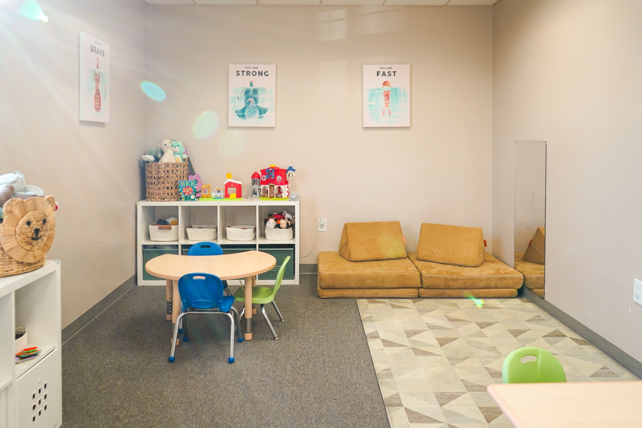 A kids' playroom with a small round table, colorful chairs, a white shelving unit filled with toys and baskets, a yellow sofa, and framed motivational posters on the walls.