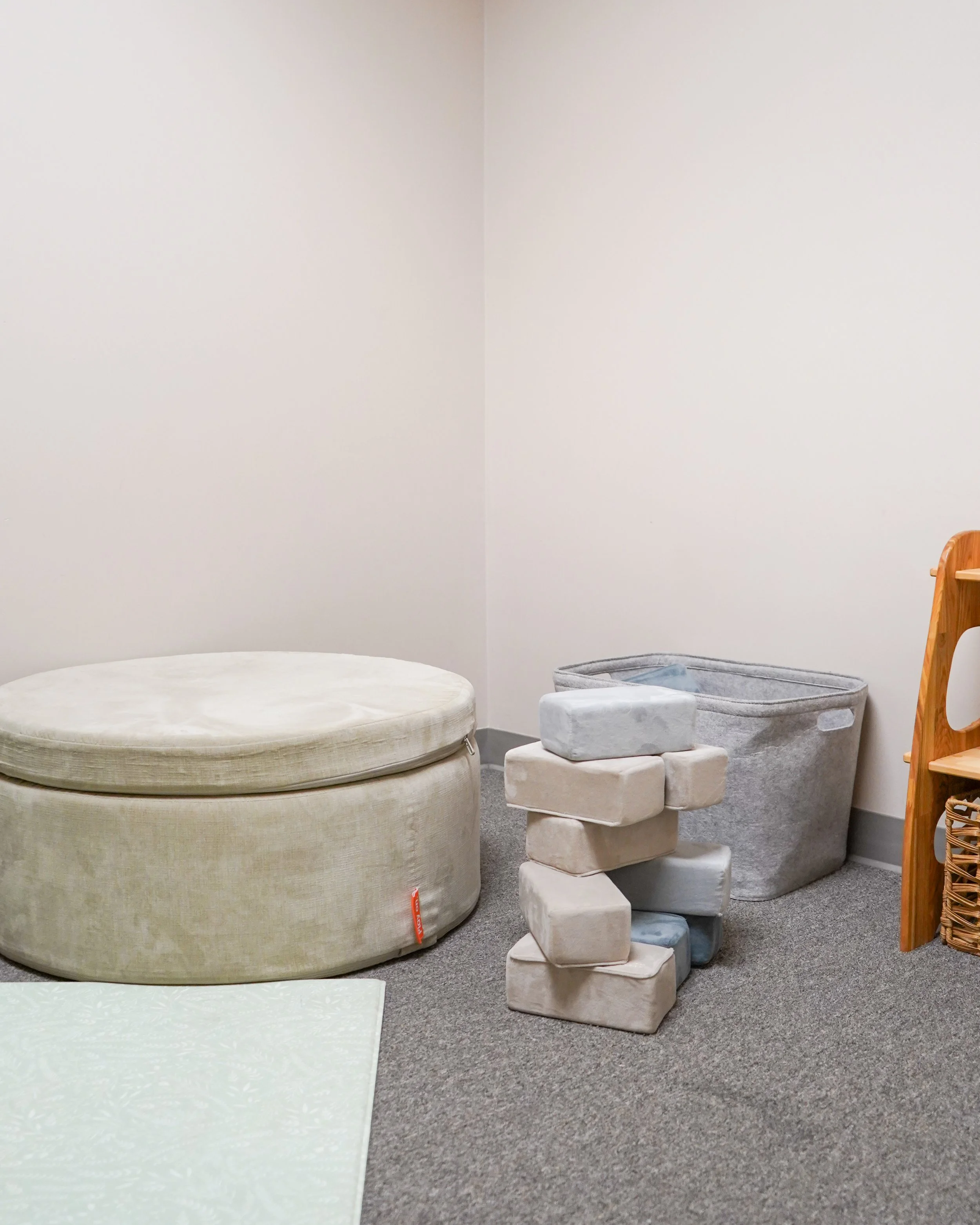 A corner of a room with a large round beige ottoman, a soft gray storage basket, a stack of soft blocks, a wooden shelf on the right, and a white wall with plain gray carpet flooring.