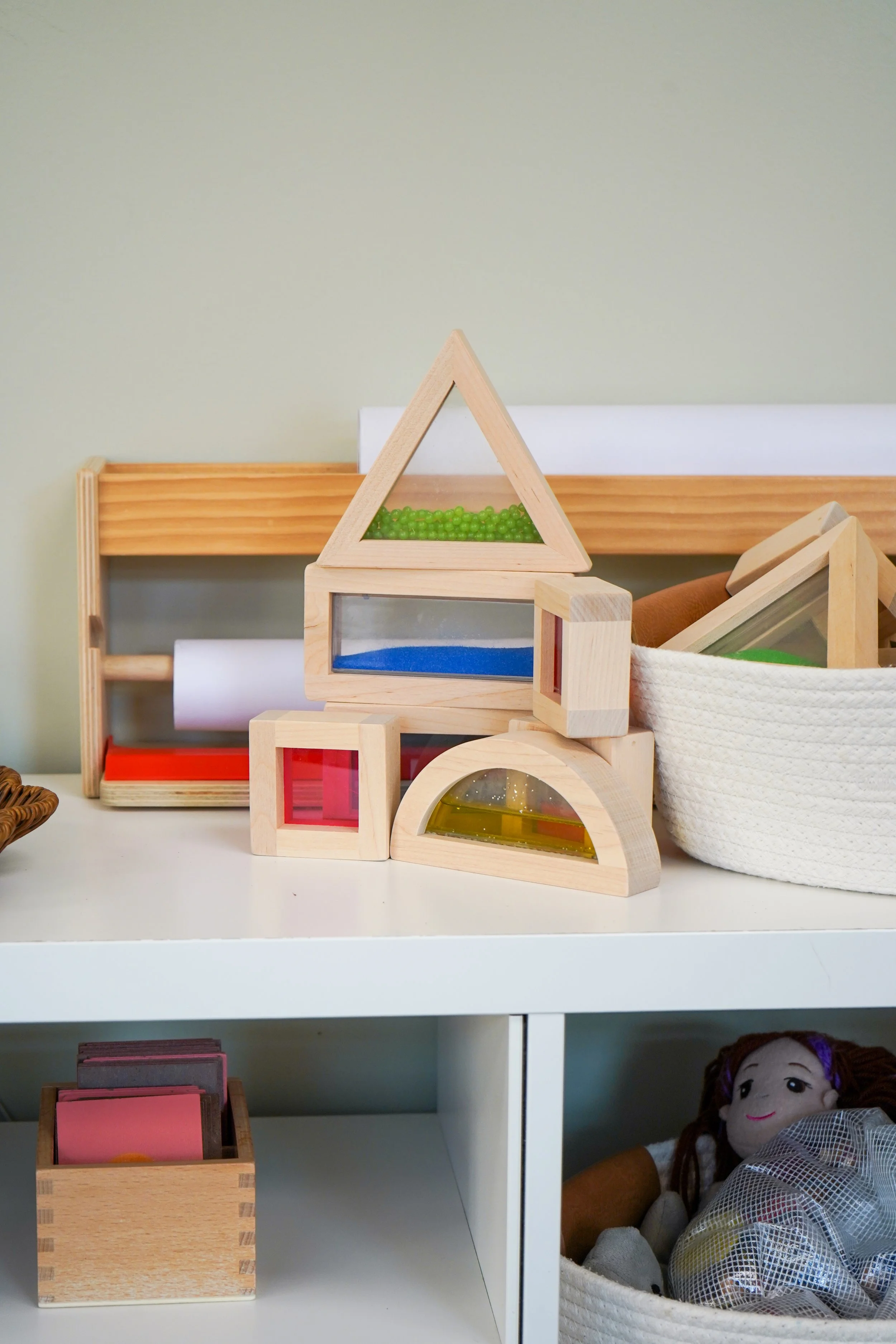 Children's toys on a white shelf, including wooden blocks with colored glass inserts, a basket with more toys, and a doll with brown hair.
