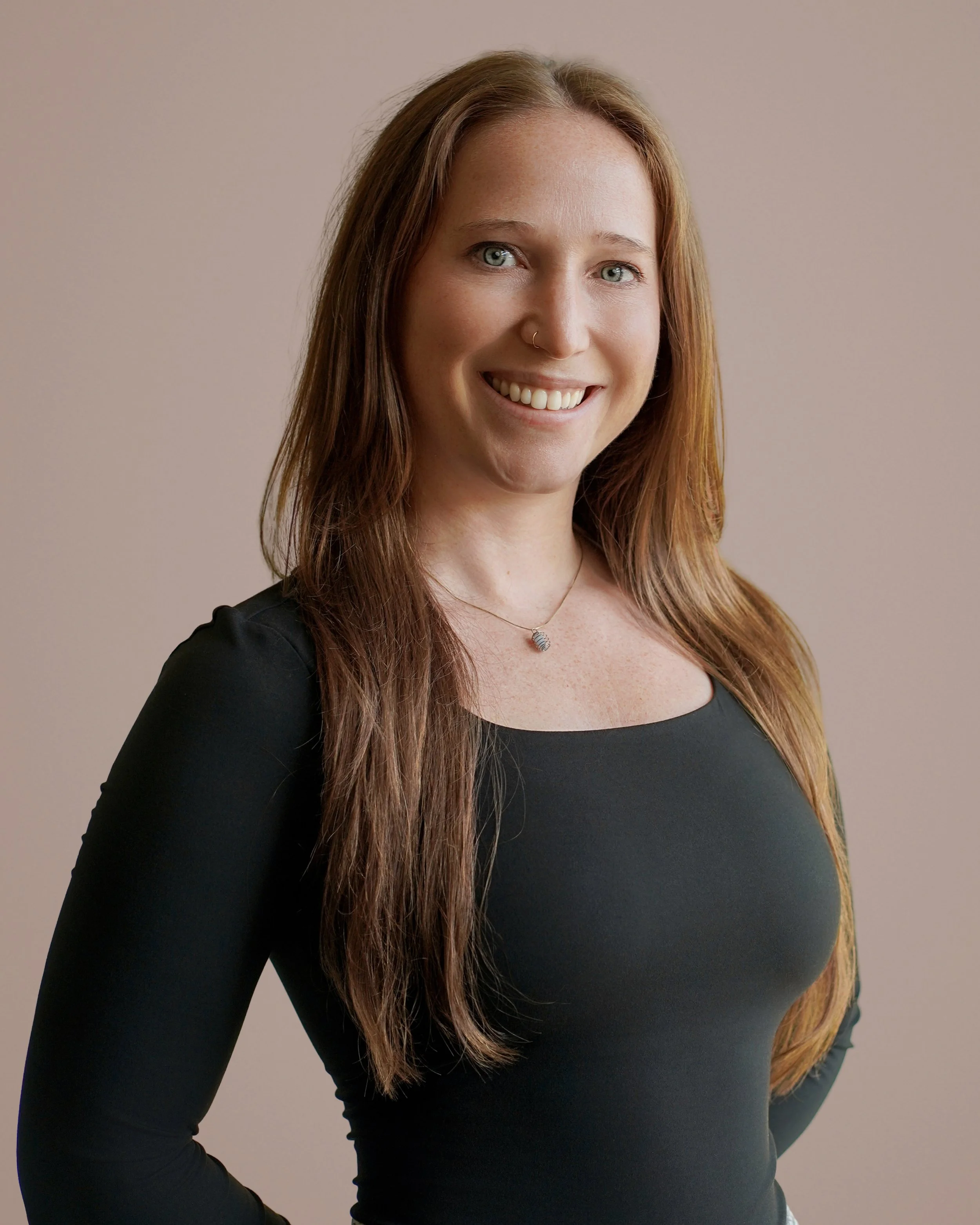A smiling woman with long red hair, wearing a black top and a necklace, standing against a plain beige background.