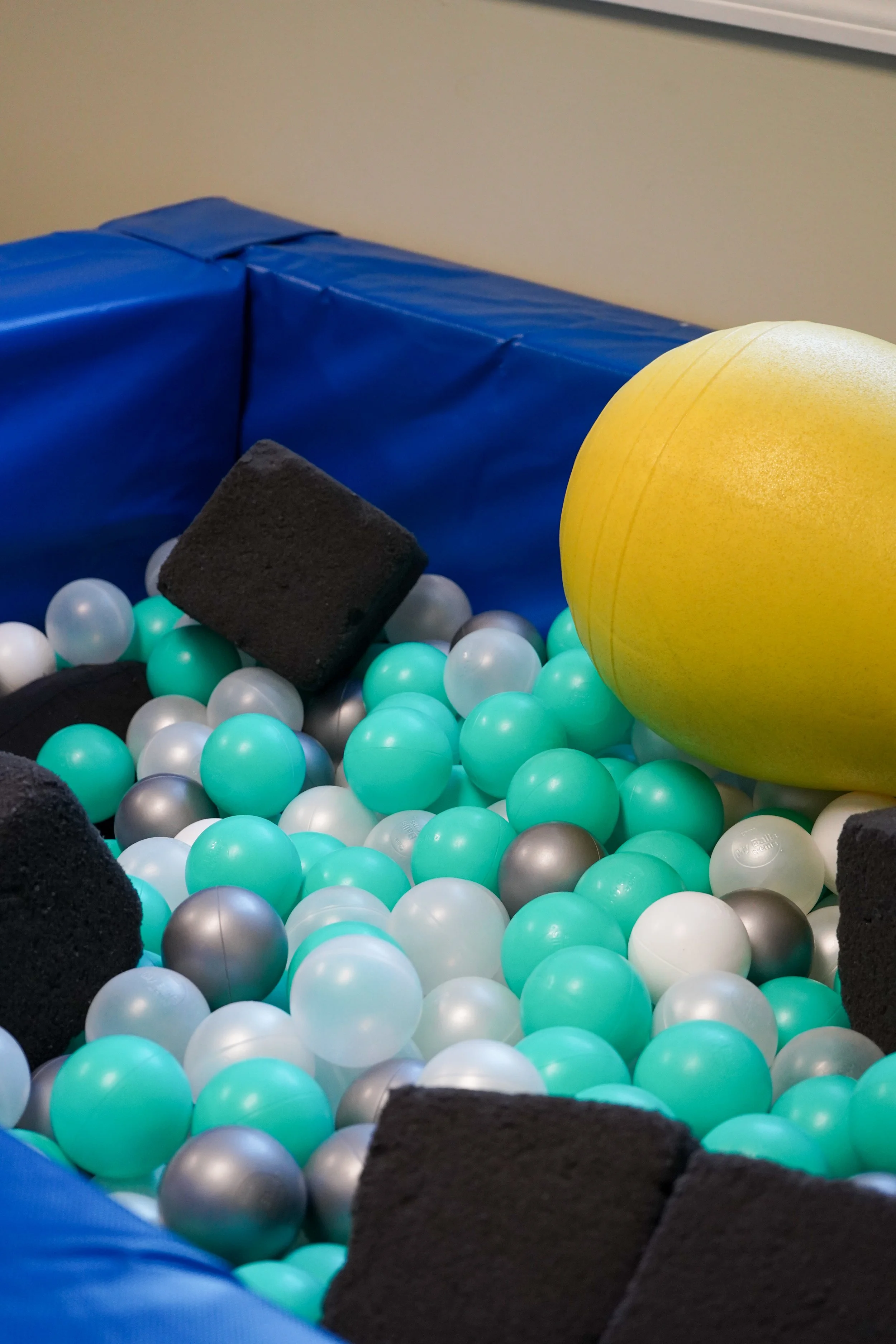 Colorful soft play area with teal, white, silver balls, large yellow ball, and black foam blocks inside a blue padded enclosure.