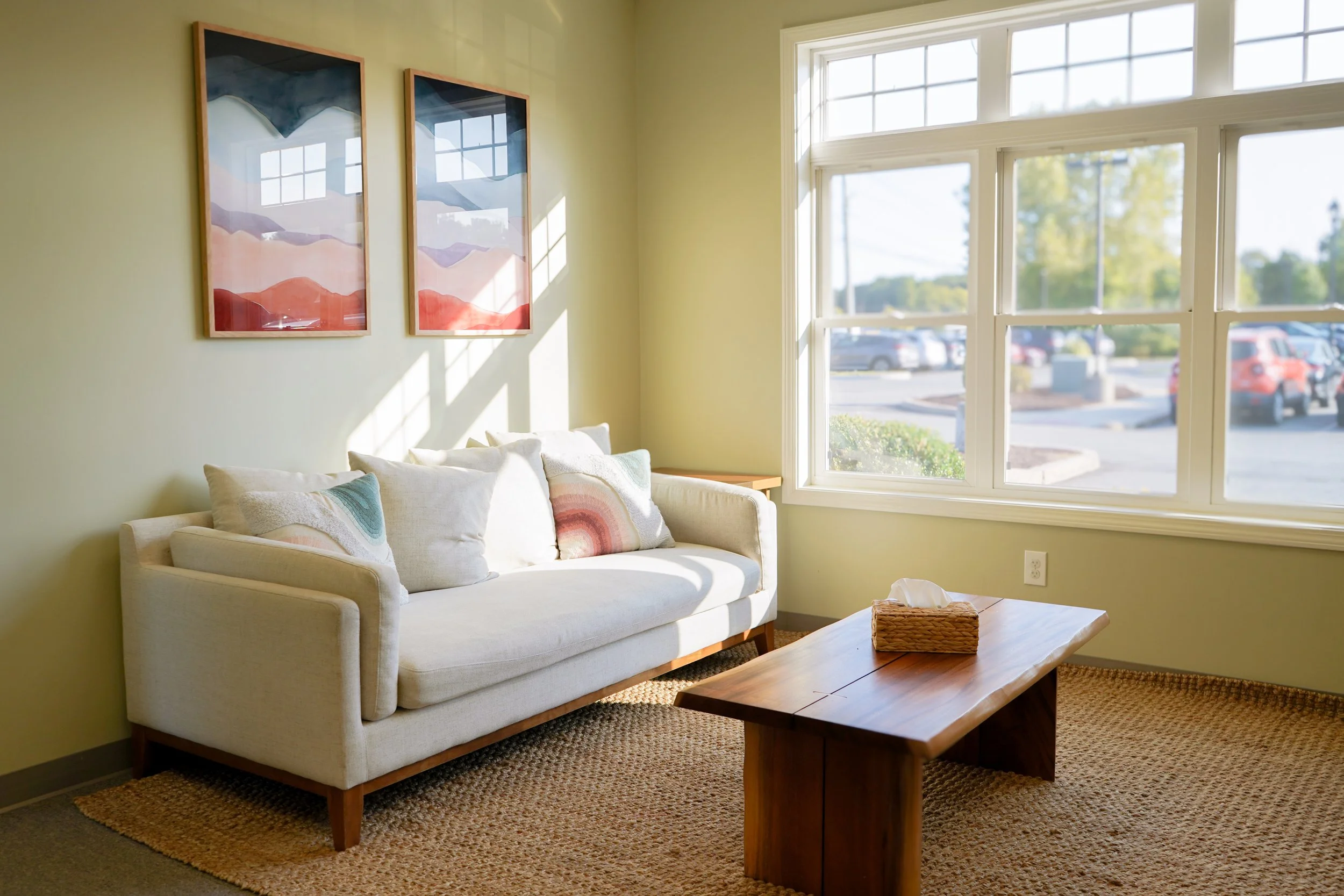 A cozy living room with a cream-colored sofa and four decorative pillows, a wooden coffee table with a tissue box, large windows allowing natural light, and art prints of colorful mountain landscapes on the wall.