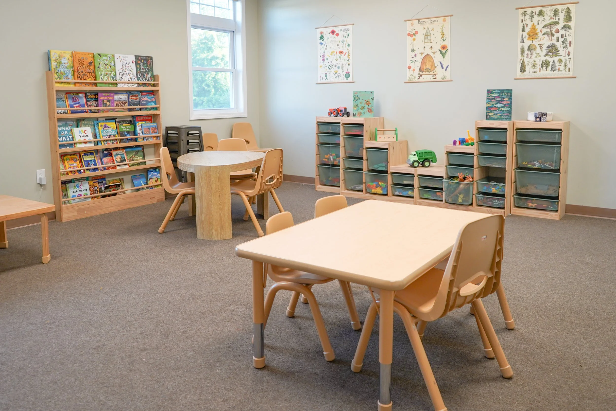 A cozy classroom with small tables and chairs, bookshelves filled with children's books, and storage units with toys and educational materials, illuminated by natural light from a window.