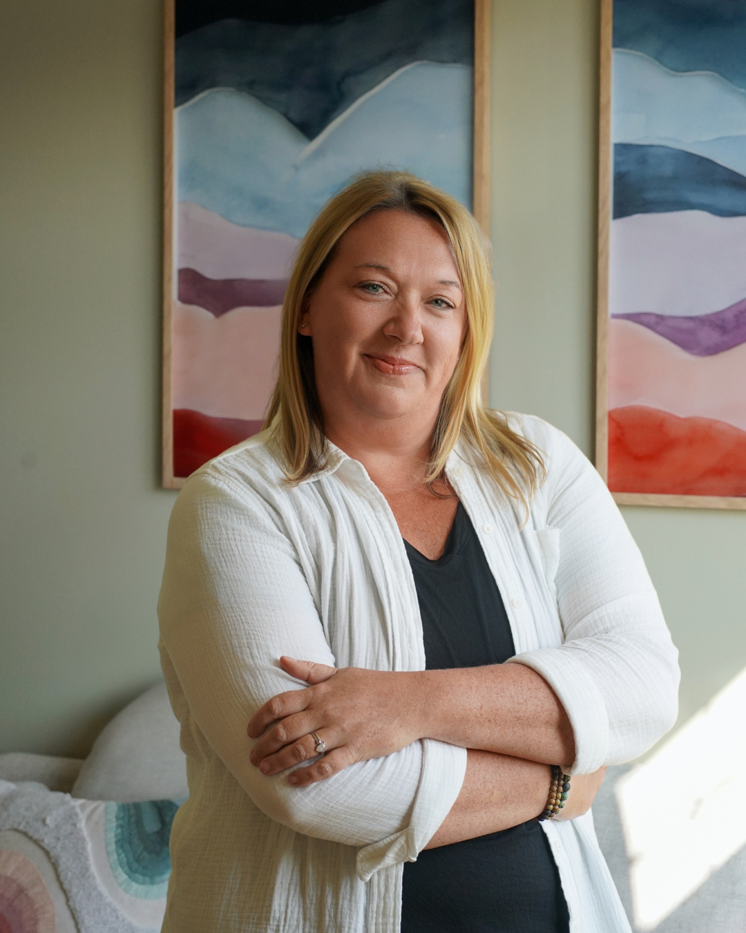 A woman with blonde hair, wearing a white shirt over a black top, standing indoors with her arms crossed and smiling. Behind her are three abstract landscape paintings with mountain and colorful sky scenes.