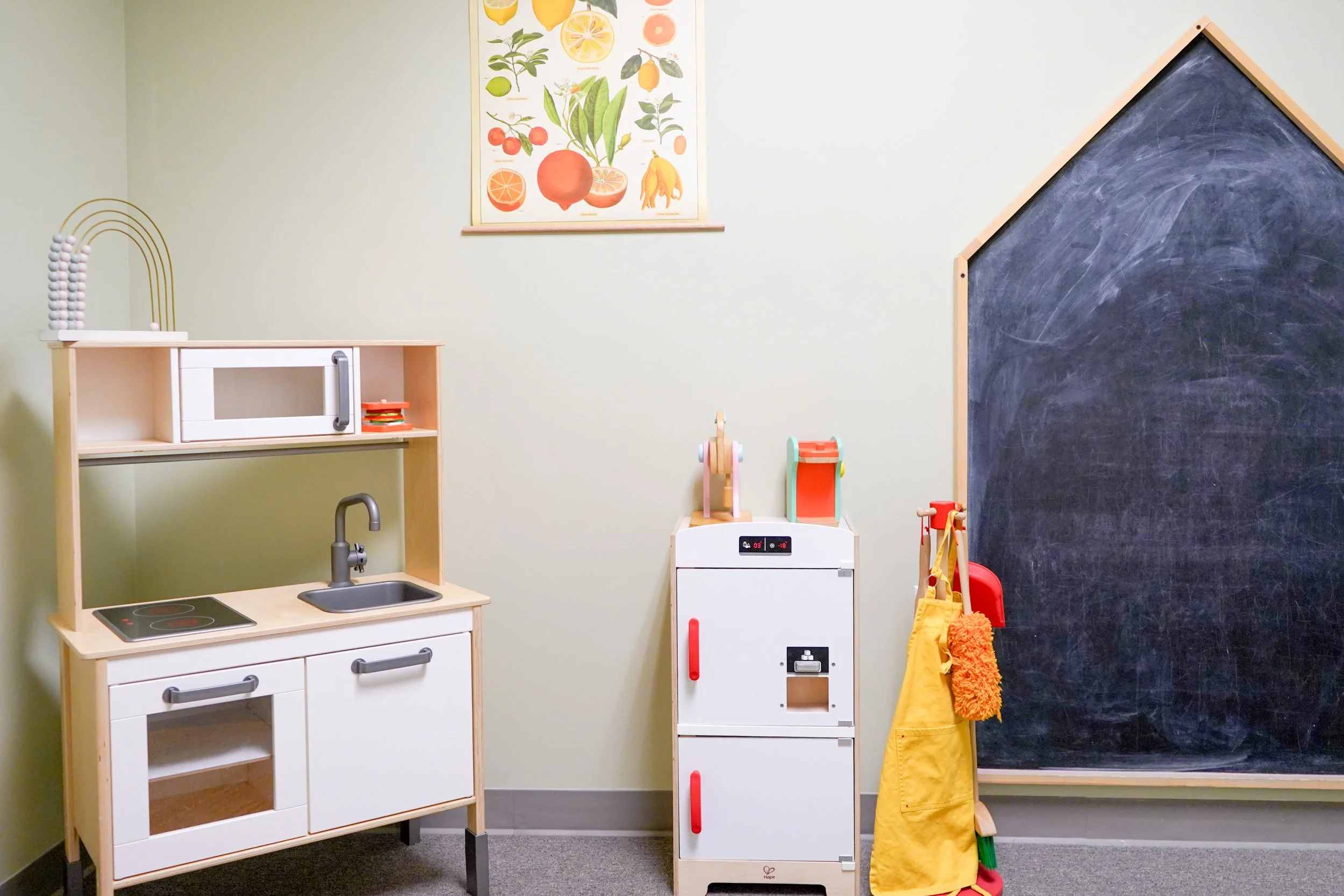 A children's play area featuring a toy kitchen set, a chalkboard wall, and various toys, with a botanical print on the wall.