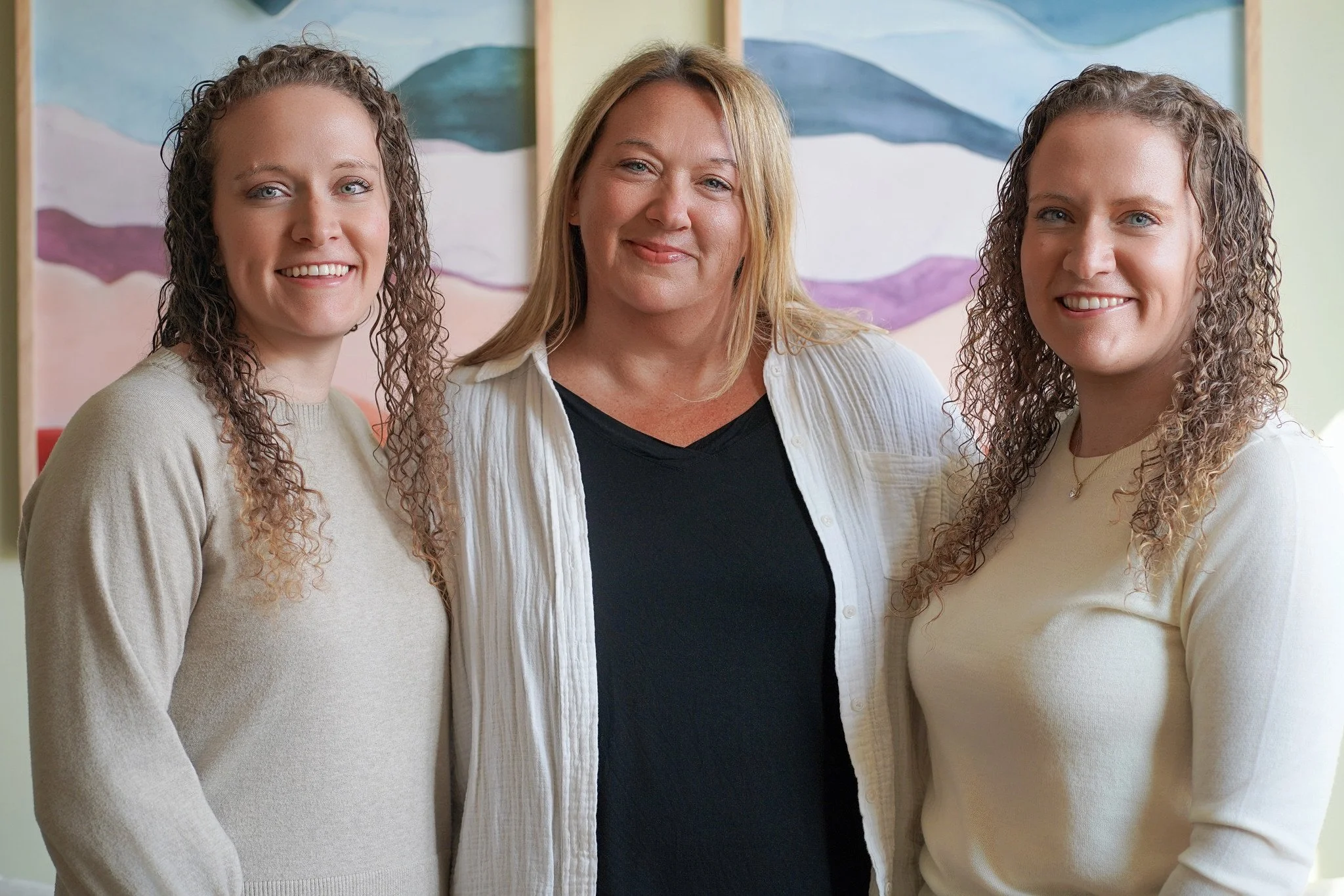 Three female founders with curly hair standing together indoors, smiling for the camera.