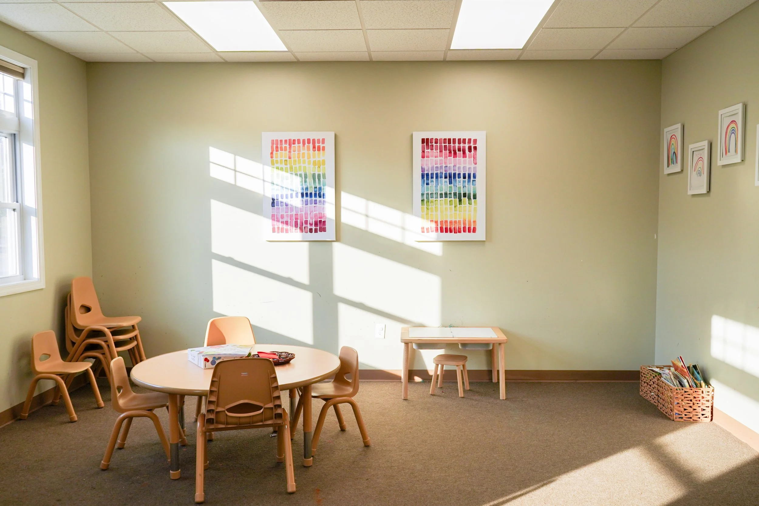 A small, empty classroom with a round table and chairs, stacked chairs in the corner, colorful artwork on the walls, and sunlight coming through the window.