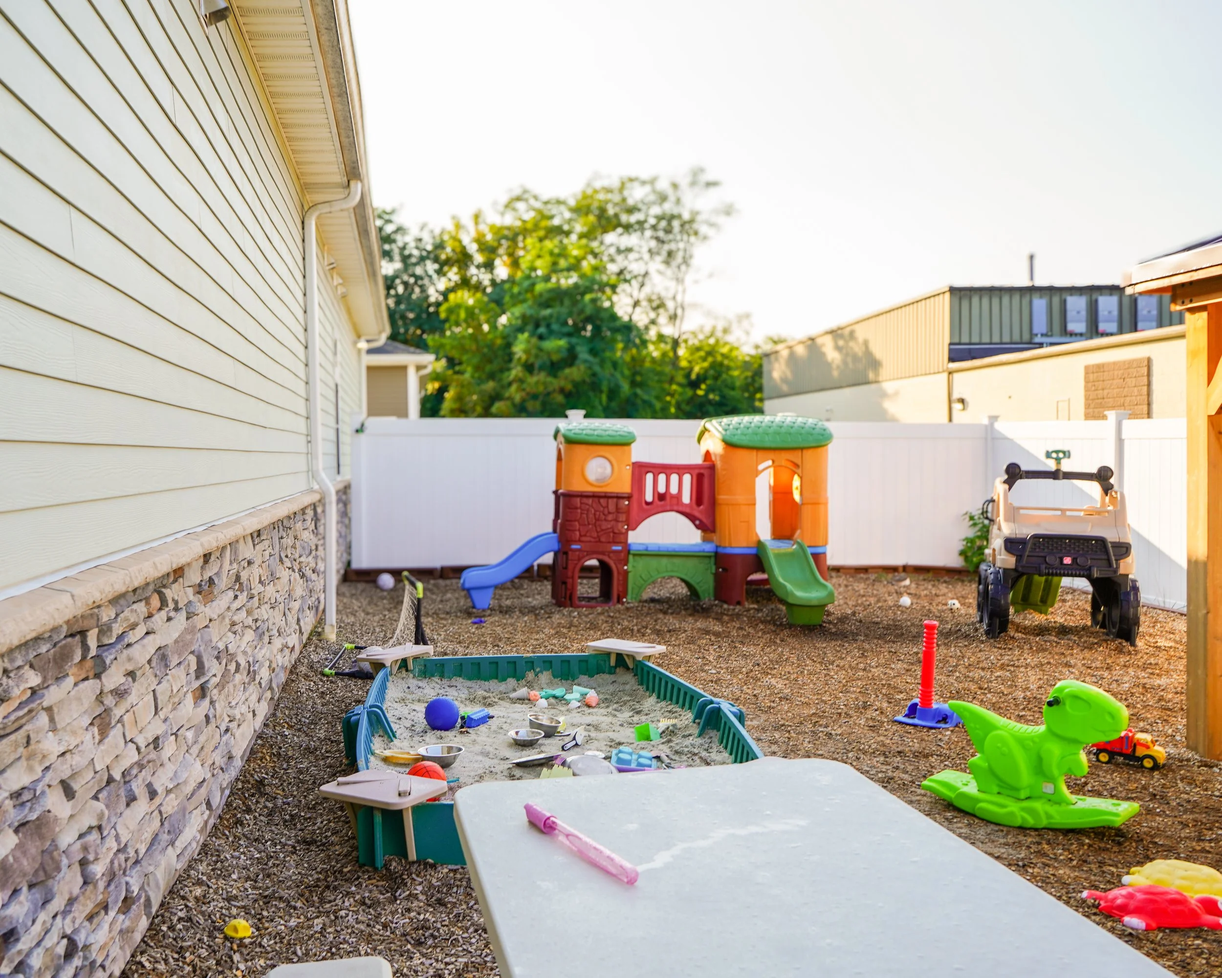 Children's backyard play area with sandpit, slides, toys, and a toy vehicle, surrounded by a white fence and located next to a house with beige siding and stone foundation.