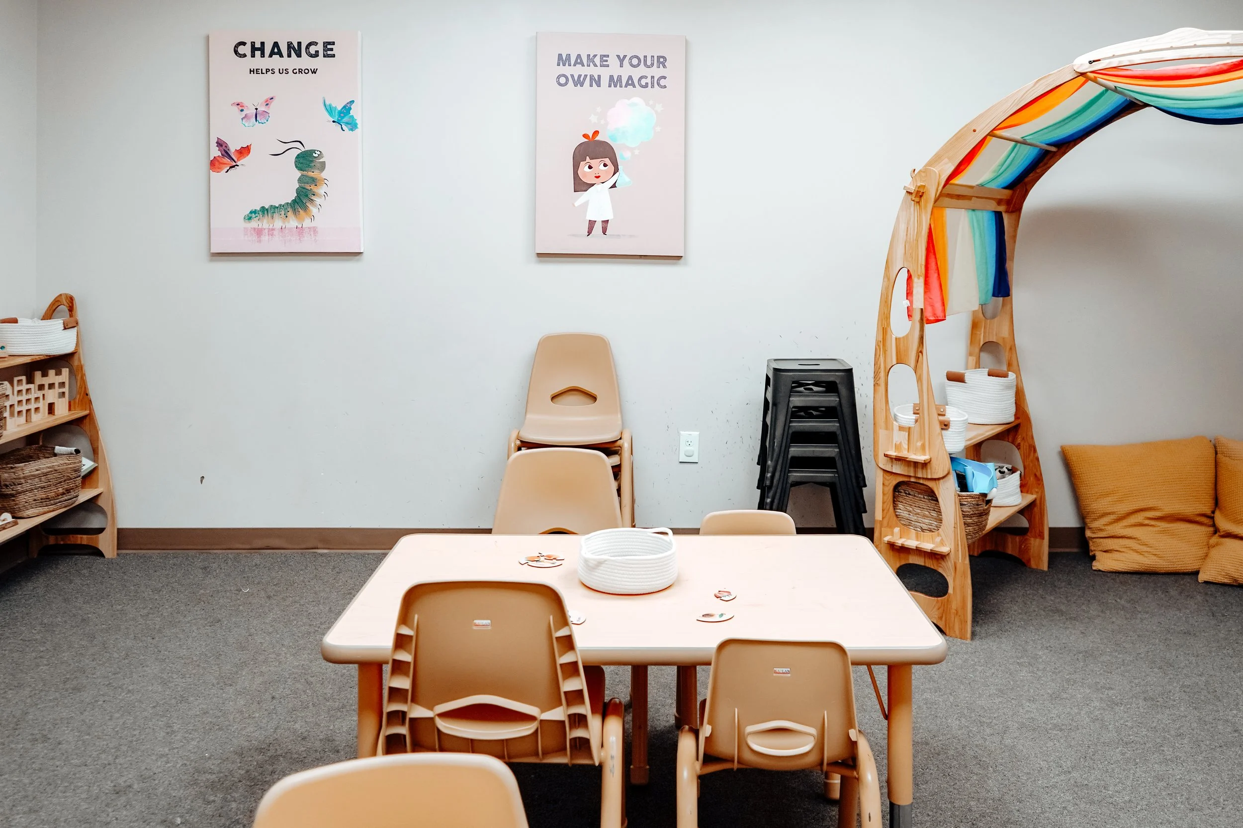 Child's playroom with a table and chairs, wall art with motivational quotes, stacked chairs, a rainbow-colored tent, shelves with baskets, and yellow cushions on the floor.