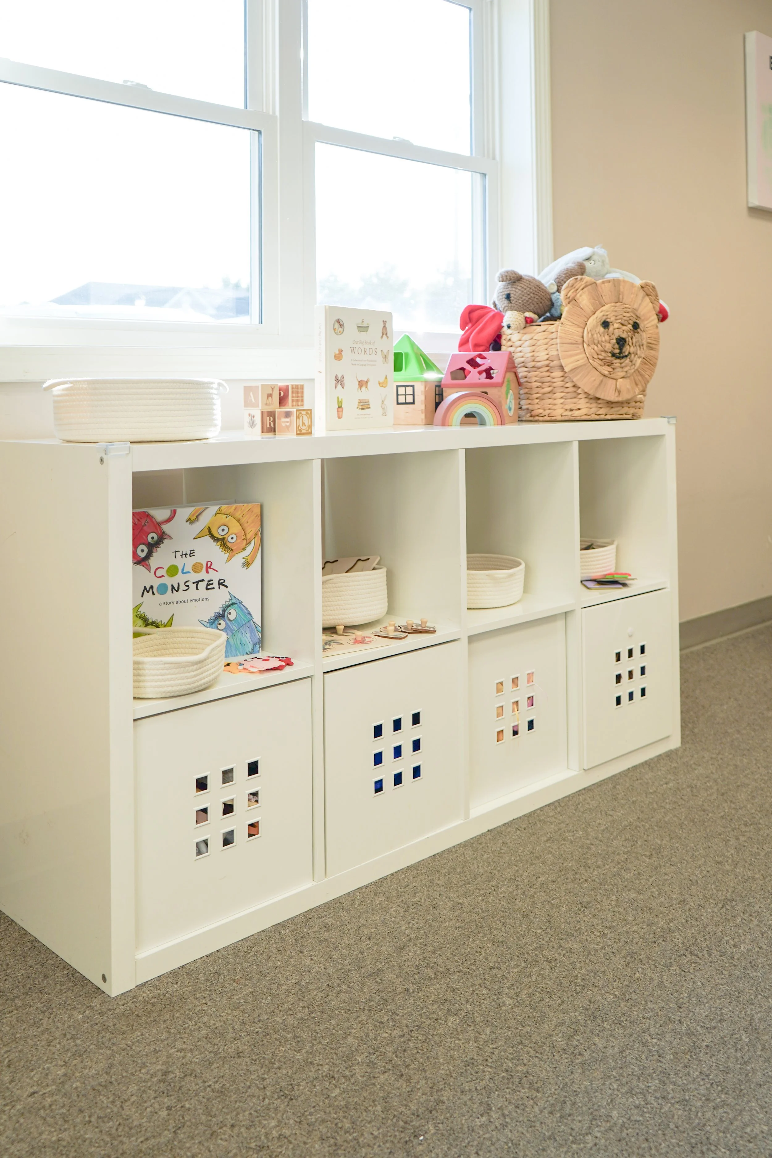 White storage unit with cubbyholes filled with baskets and toys on a beige carpet in a room with a window and beige walls.