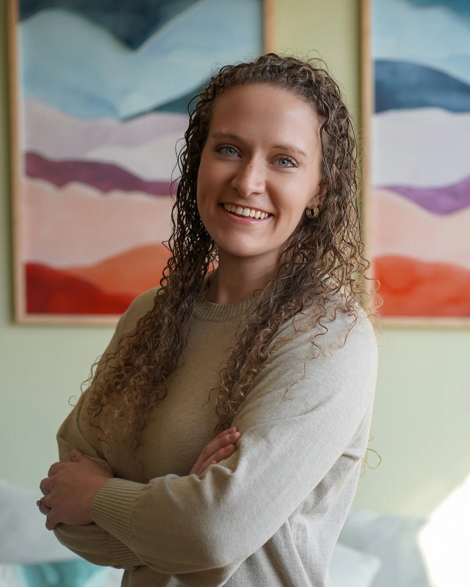 A young woman with curly brown hair and blue eyes smiling at the camera, standing in front of an abstract landscape painting in pastel colors.