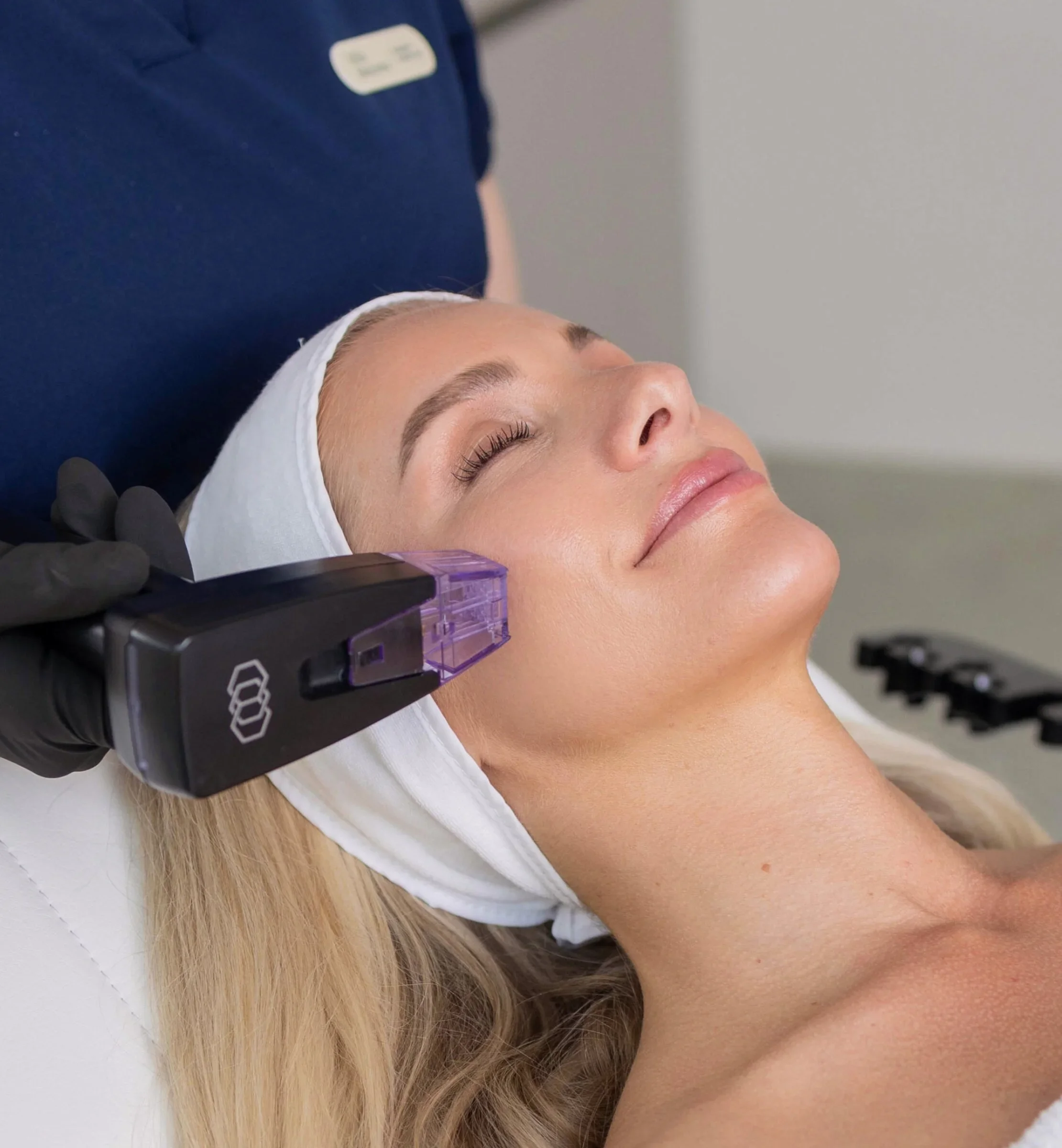 Une femme allongée, recevant un traitement facial avec un appareil de lumière, portant un bandeau blanc sur la tête, sur le visage fermé avec un léger sourire.