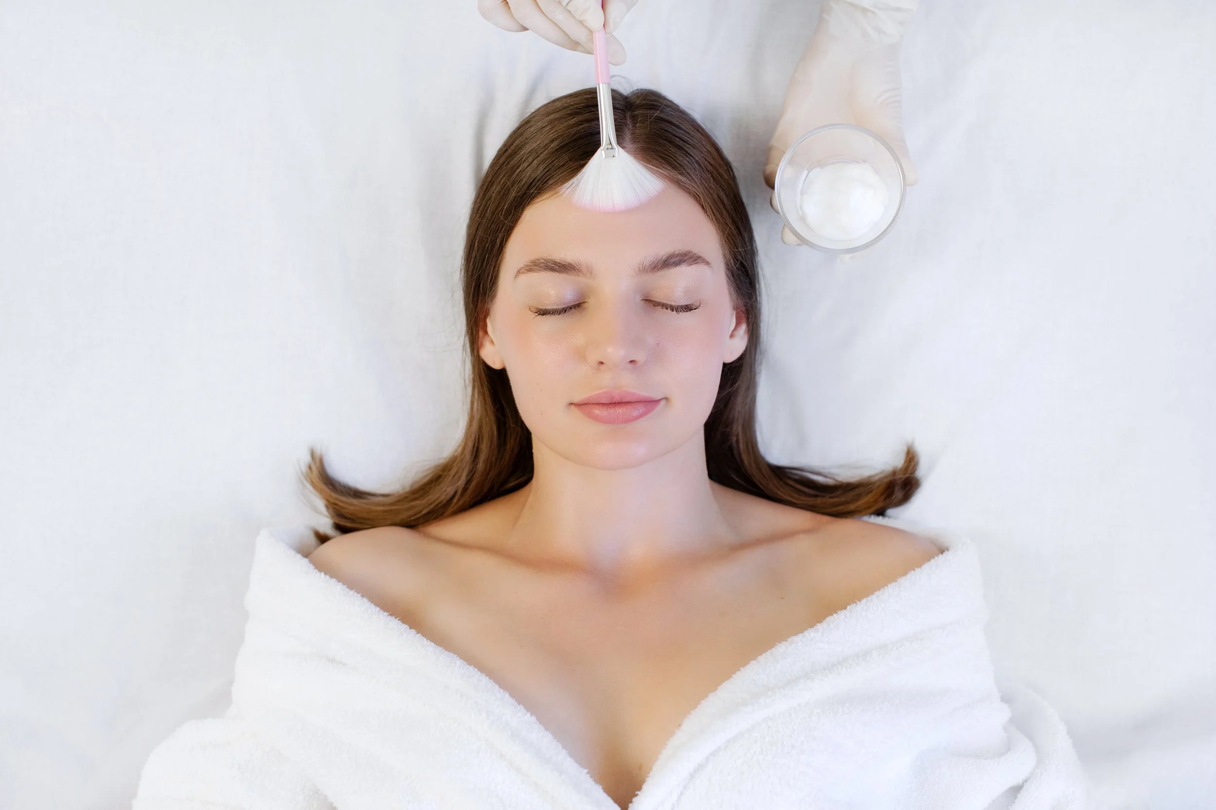 Une femme reçoit un soin du visage dans un spa, allongée sur une table, avec un chiffon blanc et un pinceau appliquant une crème sur son front.