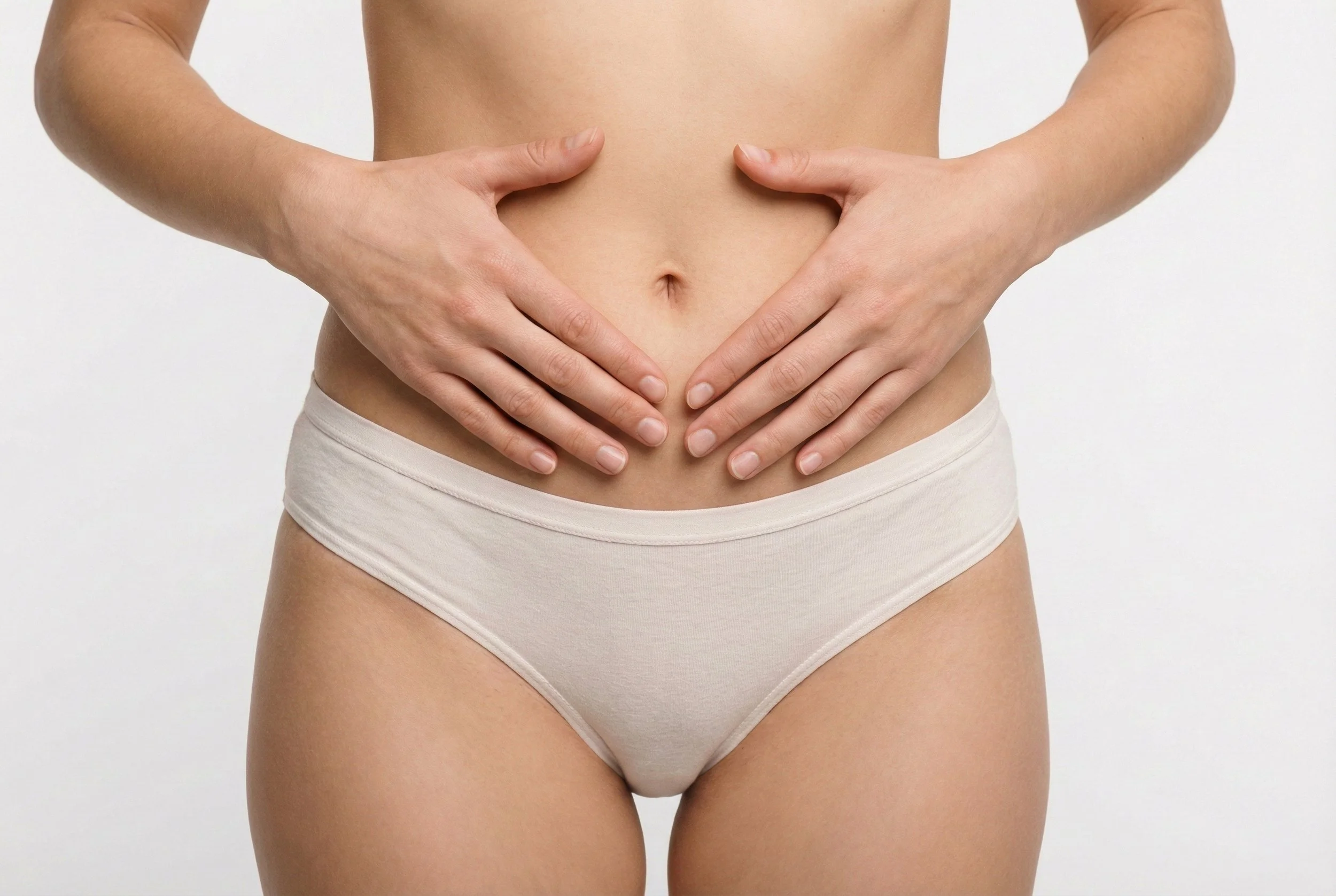 Une personne tient ses mains sur son ventre, portant des sous-vêtements beige, avec une peau claire et un ventre nu, contre un fond blanc.