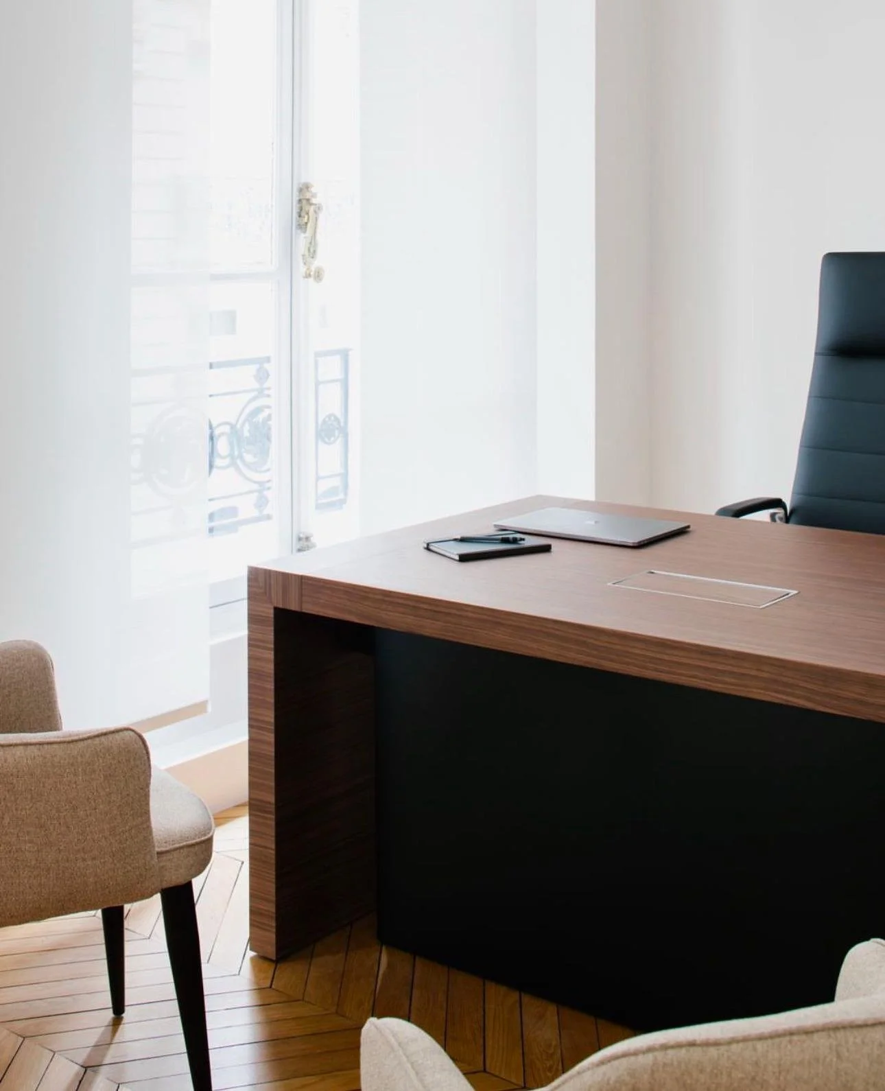 Un bureau moderne avec un fauteuil en cuir, un grand bureau en bois, une porte vitrée avec des rideaux blancs, et des chaises beige à côté.