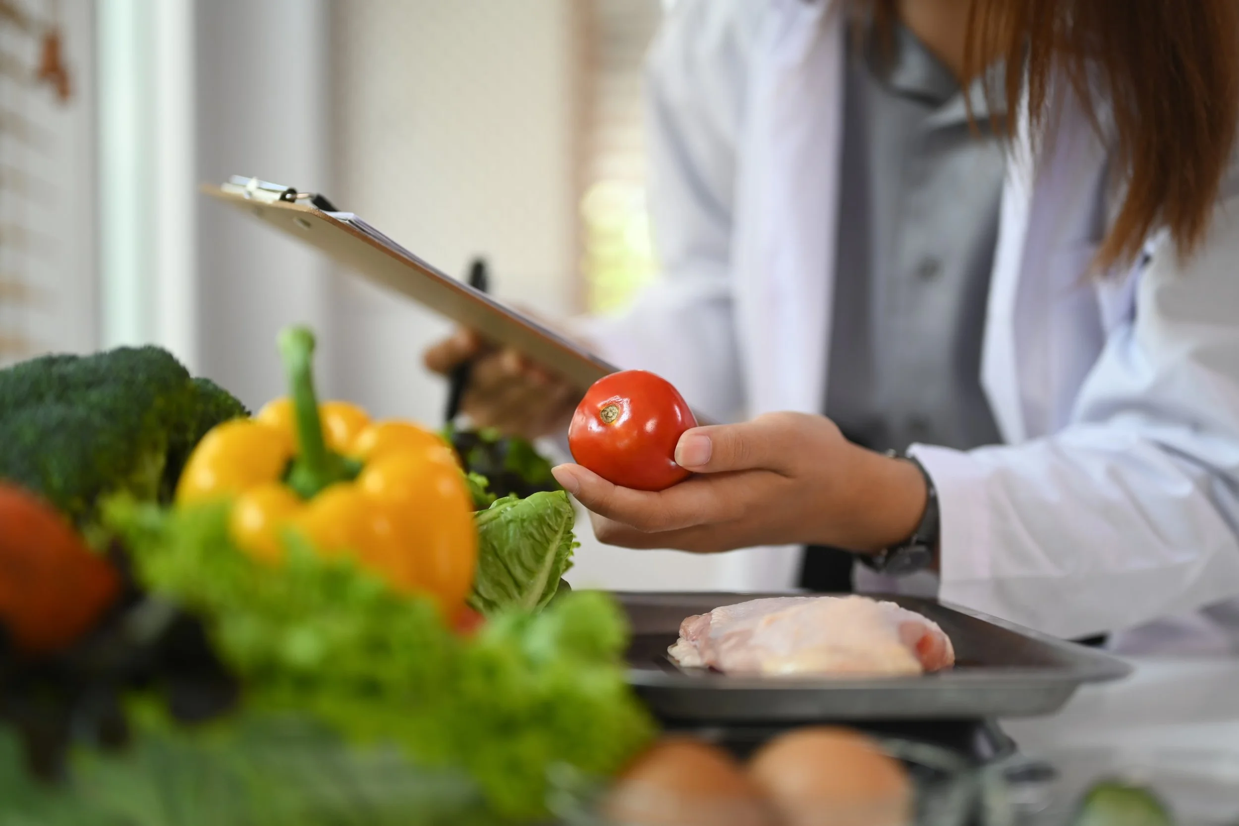 Une personne en blouse blanche examine une tomate rouge dans une cuisine, entourée de légumes frais, y compris un brocoli, un poivron jaune, des feuilles de laitue, et des œufs.