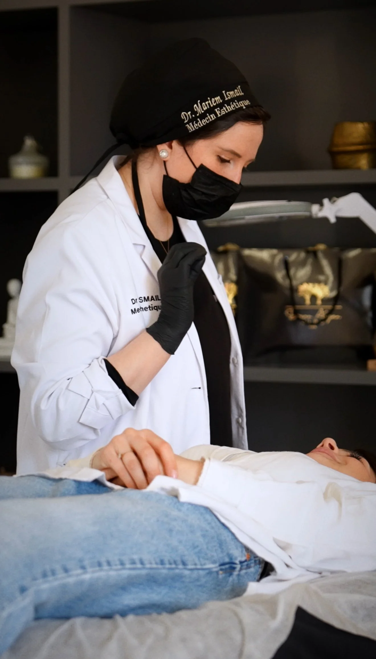 Une femme en blouse blanche, portant un masque noir et un foulard noir avec inscription, semble administrer un traitement à une personne allongée sur une table. Elle porte des gants noirs et elle est dans un environnement professionnel, probablement une clinique ou un cabinet médical.