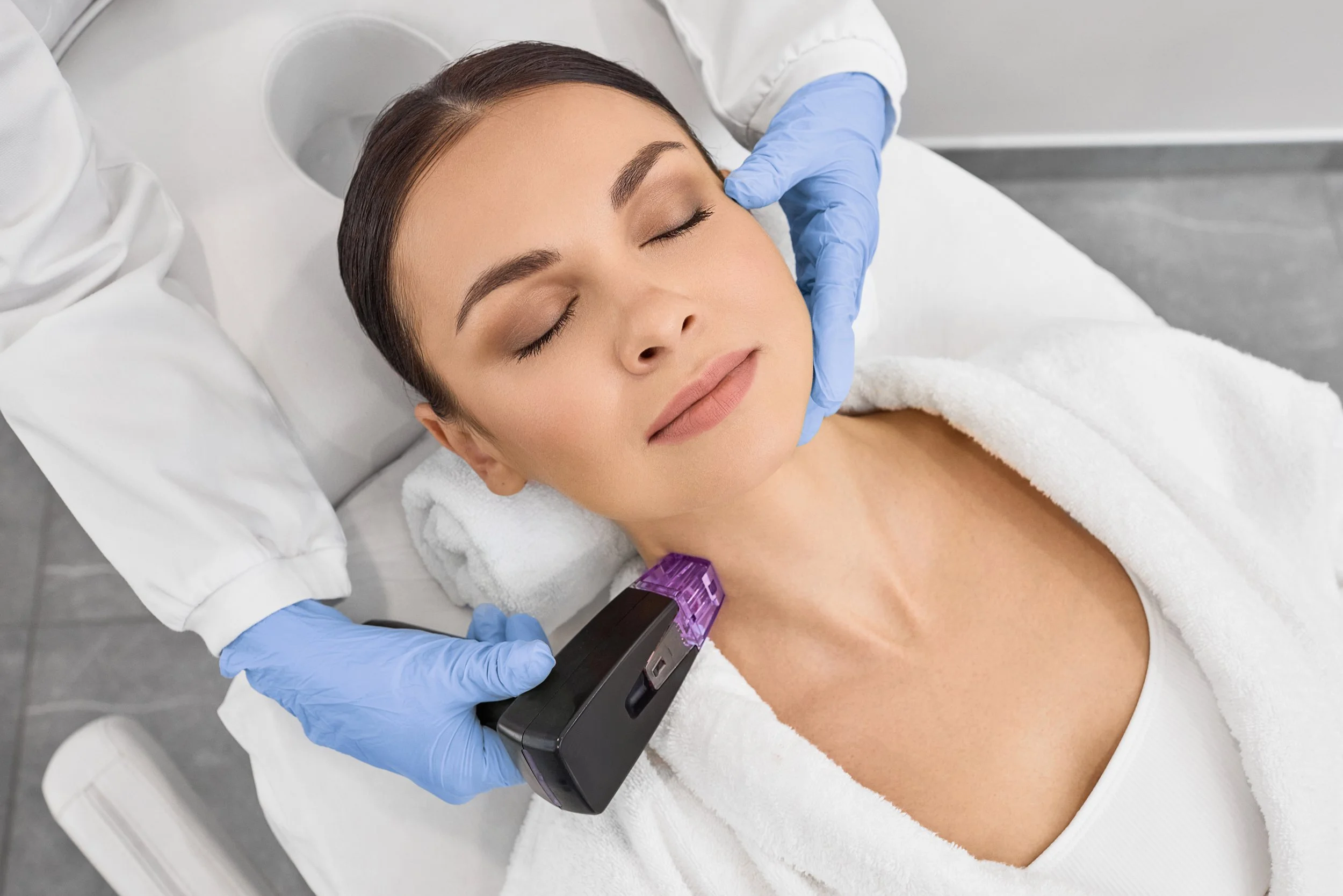 Une femme reçoit un traitement de beauté au visage, organisée par un professionnel en laboratoire esthétique, avec un appareil de lumière placé sur le cou.