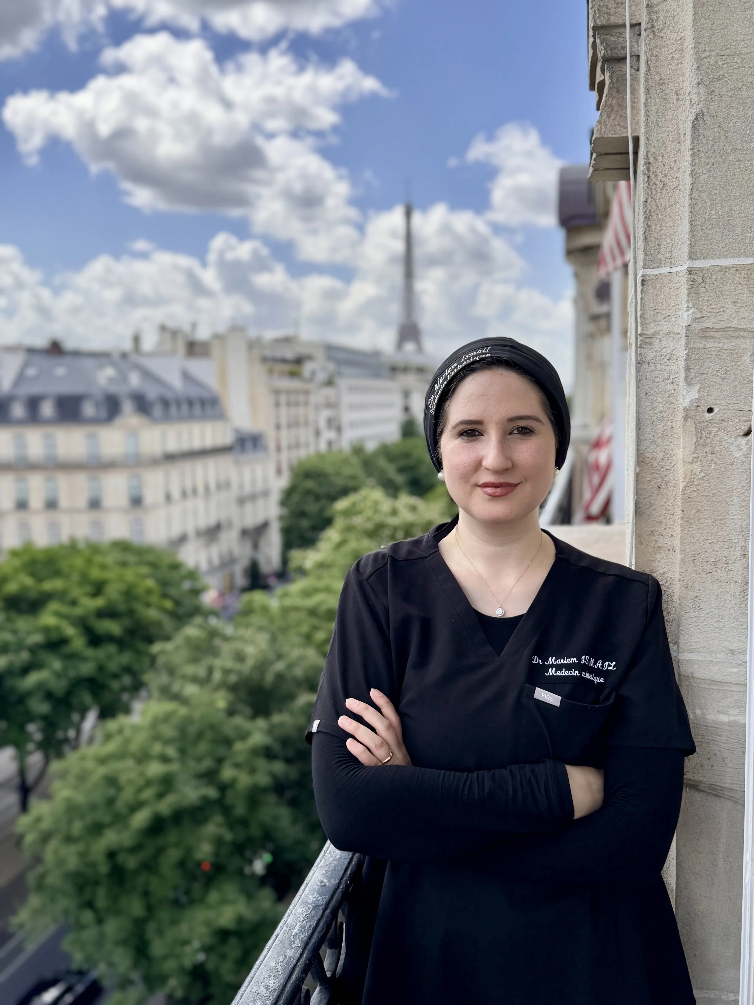 Une femme portant une blouse noire avec un nom brodé, debout sur un balcon avec vue sur la ville de Paris, avec la Tour Eiffel en arrière-plan, par une journée ensoleillée avec des nuages blancs dans le ciel.