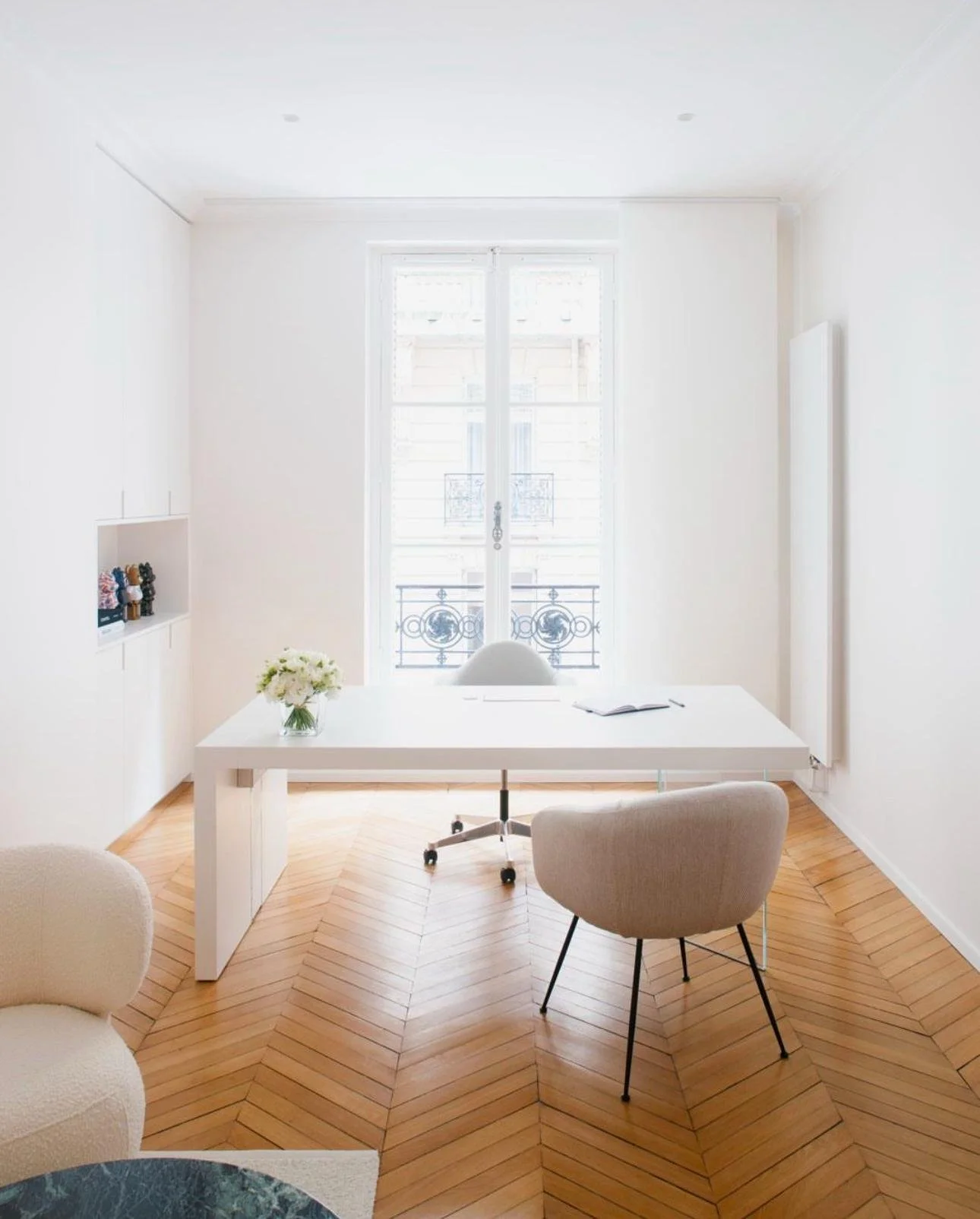 Salon minimaliste avec table blanche, deux chaises beige, parquet en bois, grande fenêtre avec balcon.