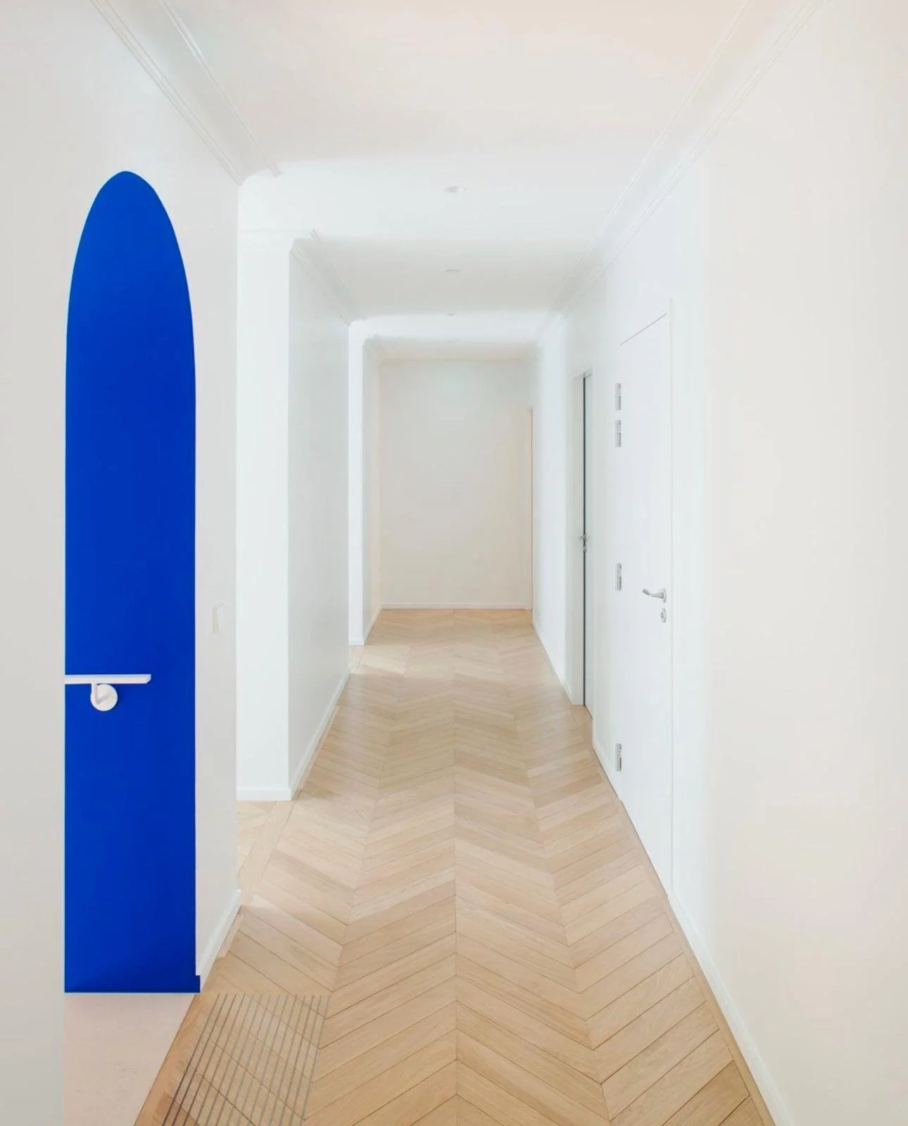 Couloir blanc avec porte blanche, sol en bois clair en motif chevrons, et une grande illustration de forme arrondie bleue sur le mur gauche.