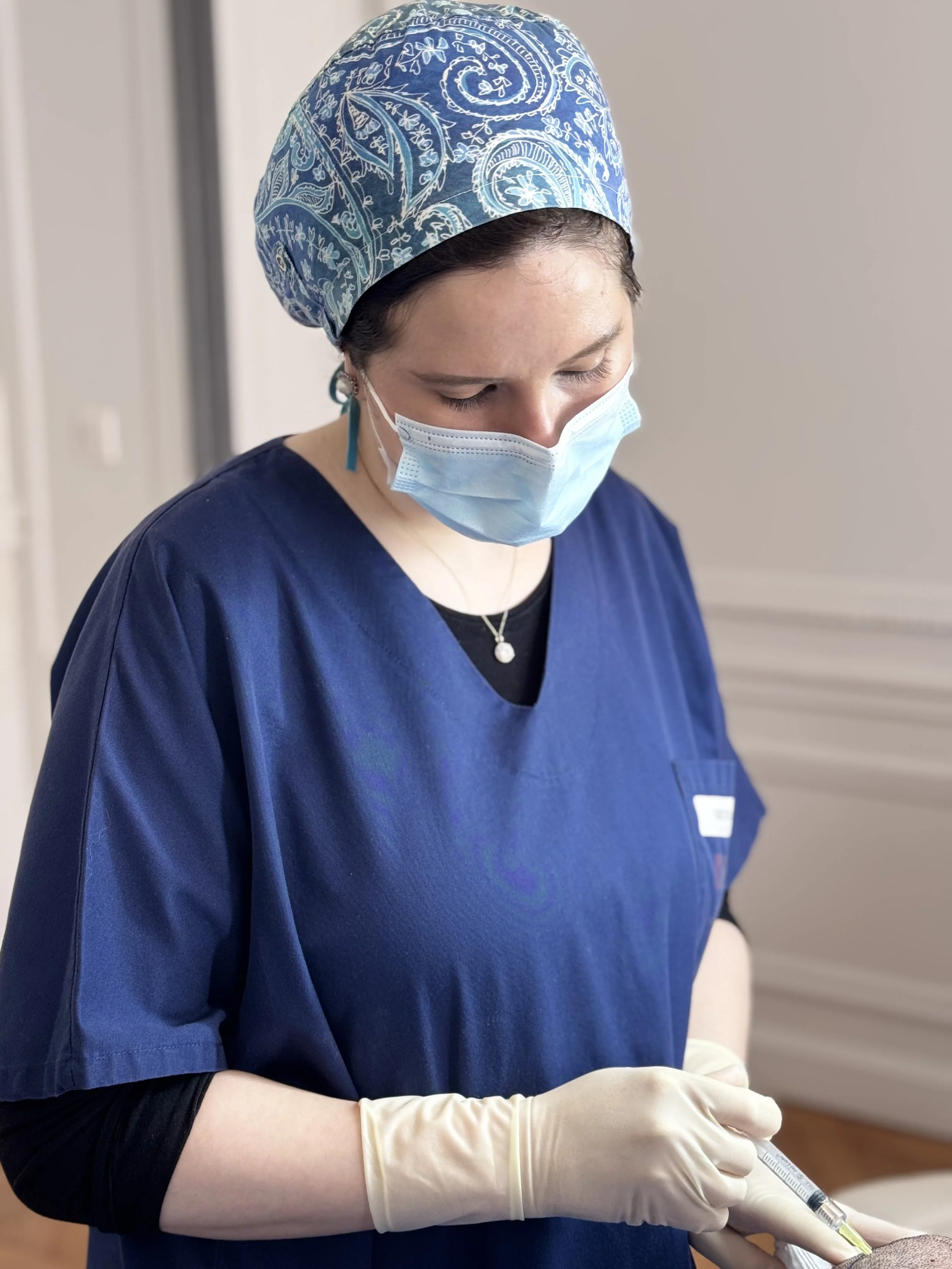 Une infirmière ou une professionnelle de la santé portant une coiffe bleue à motifs, un masque chirurgical, une blouse bleue et des gants en latex beige, préparant une injection ou une prise de sang.