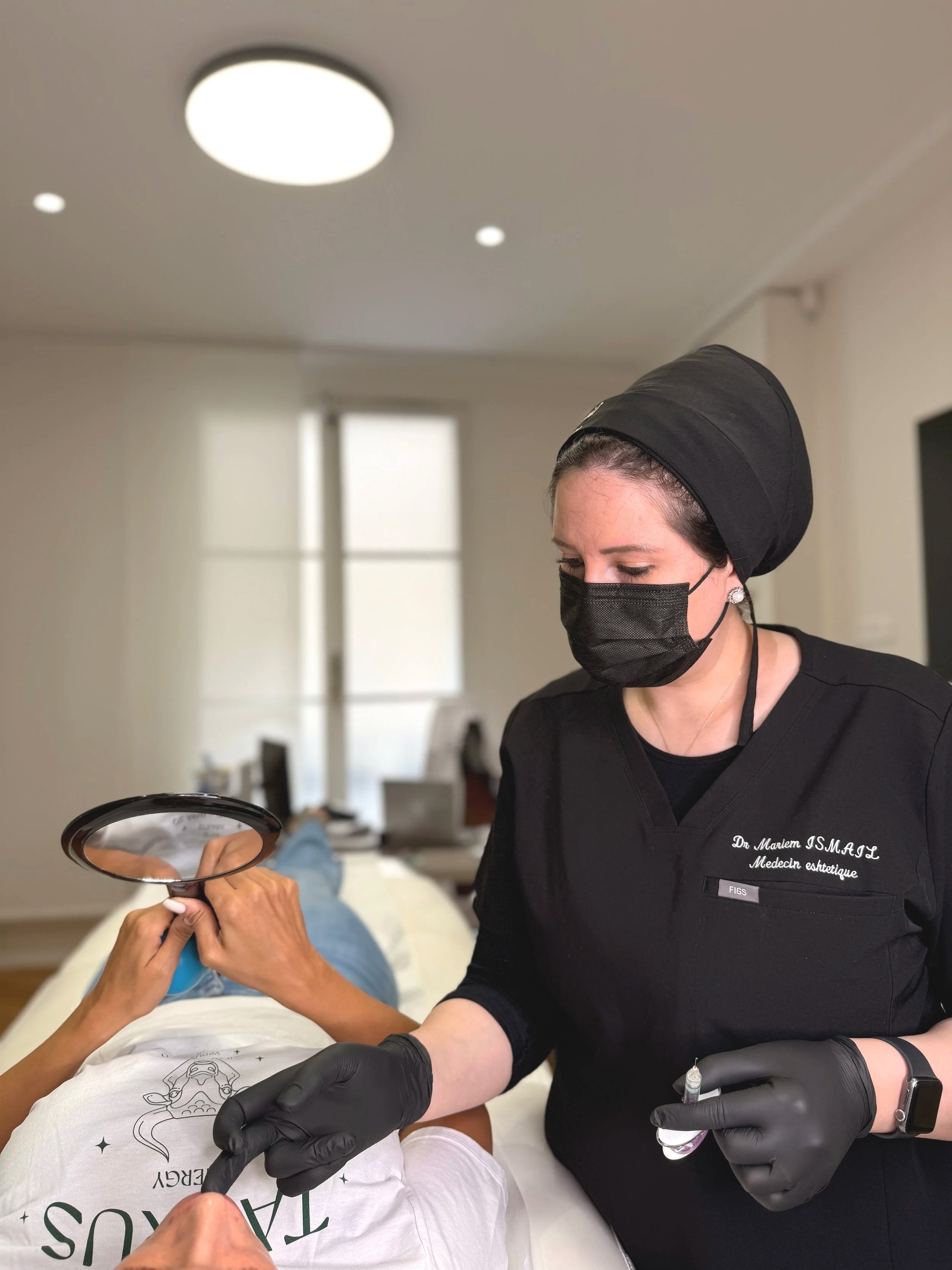 Une femme à l'uniforme médical, portant un masque et des gants noirs, administrant une injection à une personne allongée sur un lit d'examen. La femme porte une coiffe noire et une badge avec le nom et le titre.