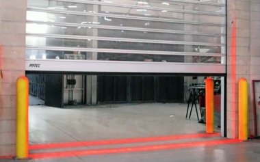 A partially open parking garage door revealing an empty indoor parking area with orange safety bollards and Rytec Pathwatch lights.