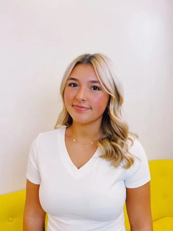 A young woman with blonde hair and a white V-neck T-shirt sitting on a yellow sofa against a plain beige wall.