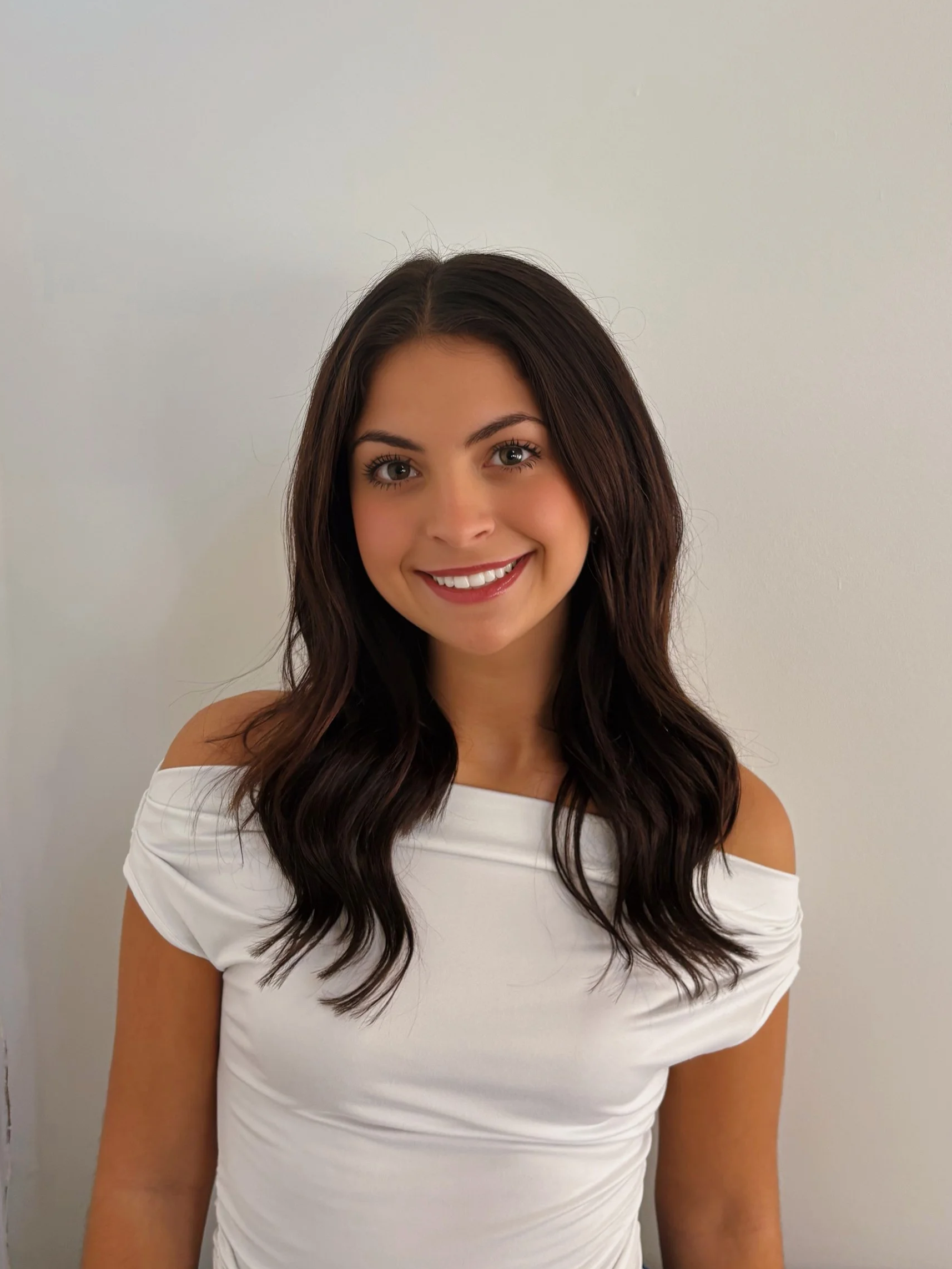 Portrait of a smiling young woman with long, wavy dark hair wearing a white off-shoulder top, standing against a plain light-colored wall.