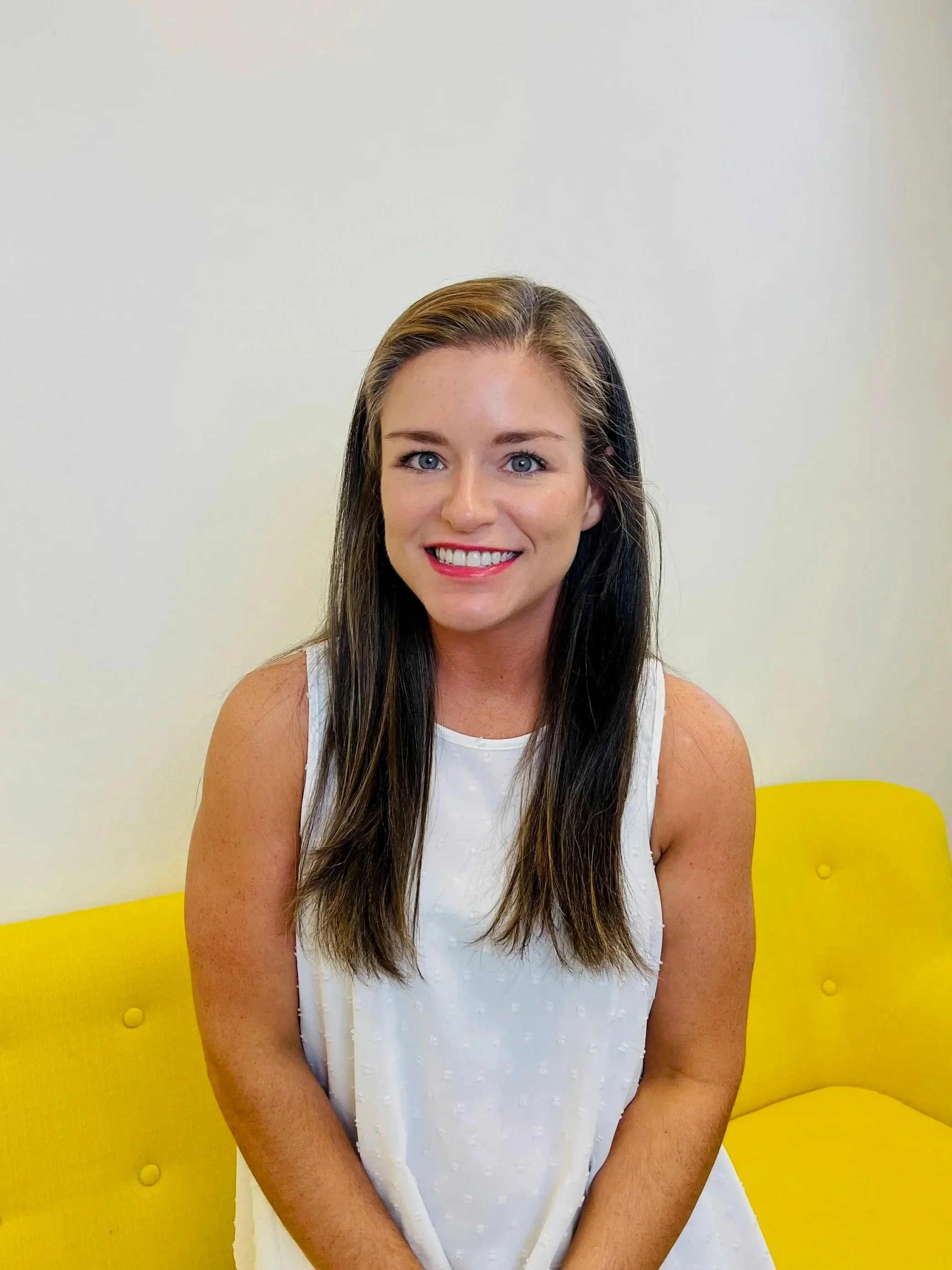 A young woman with long brown hair and blue eyes, smiling, wearing a sleeveless white top, sitting on a yellow sofa against a plain white wall.