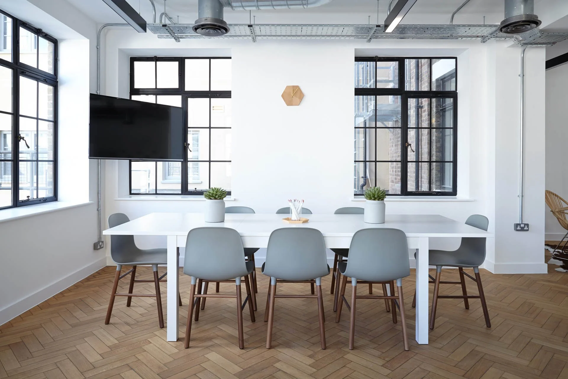A modern conference room with a long white table, nine gray chairs, and two potted plants. Large windows with black frames let in natural light, and a flat-screen TV is mounted on the wall.