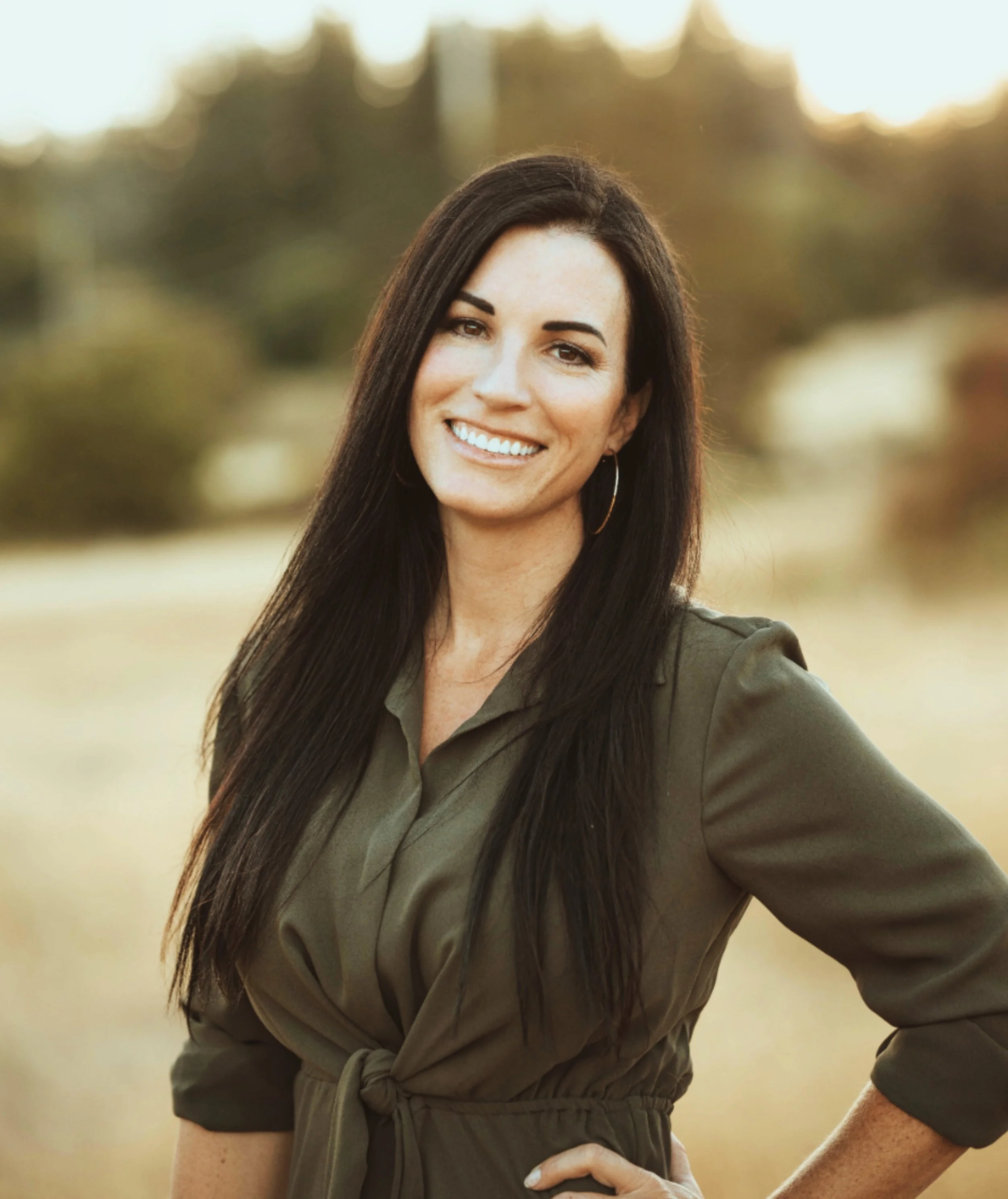 A woman with long dark hair smiling outdoors in a field, wearing an olive green dress with three-quarter sleeves and hoop earrings.