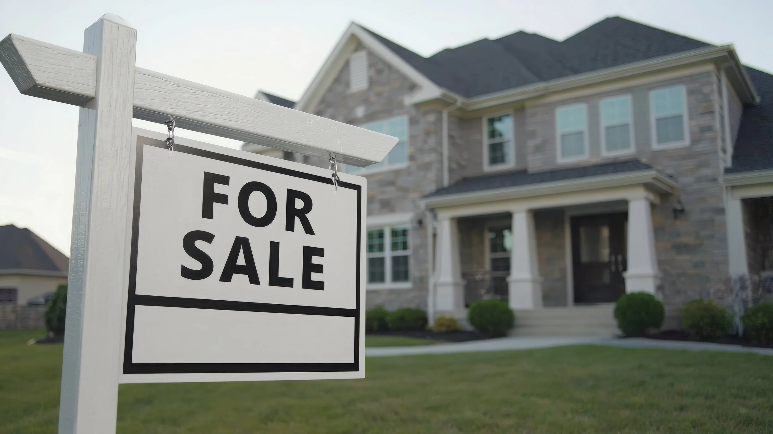 A white sign with black letters that reads "For Sale" is posted in front of a large, modern house with a brick and stone exterior, several windows, a porch with columns, and a well-manicured lawn.