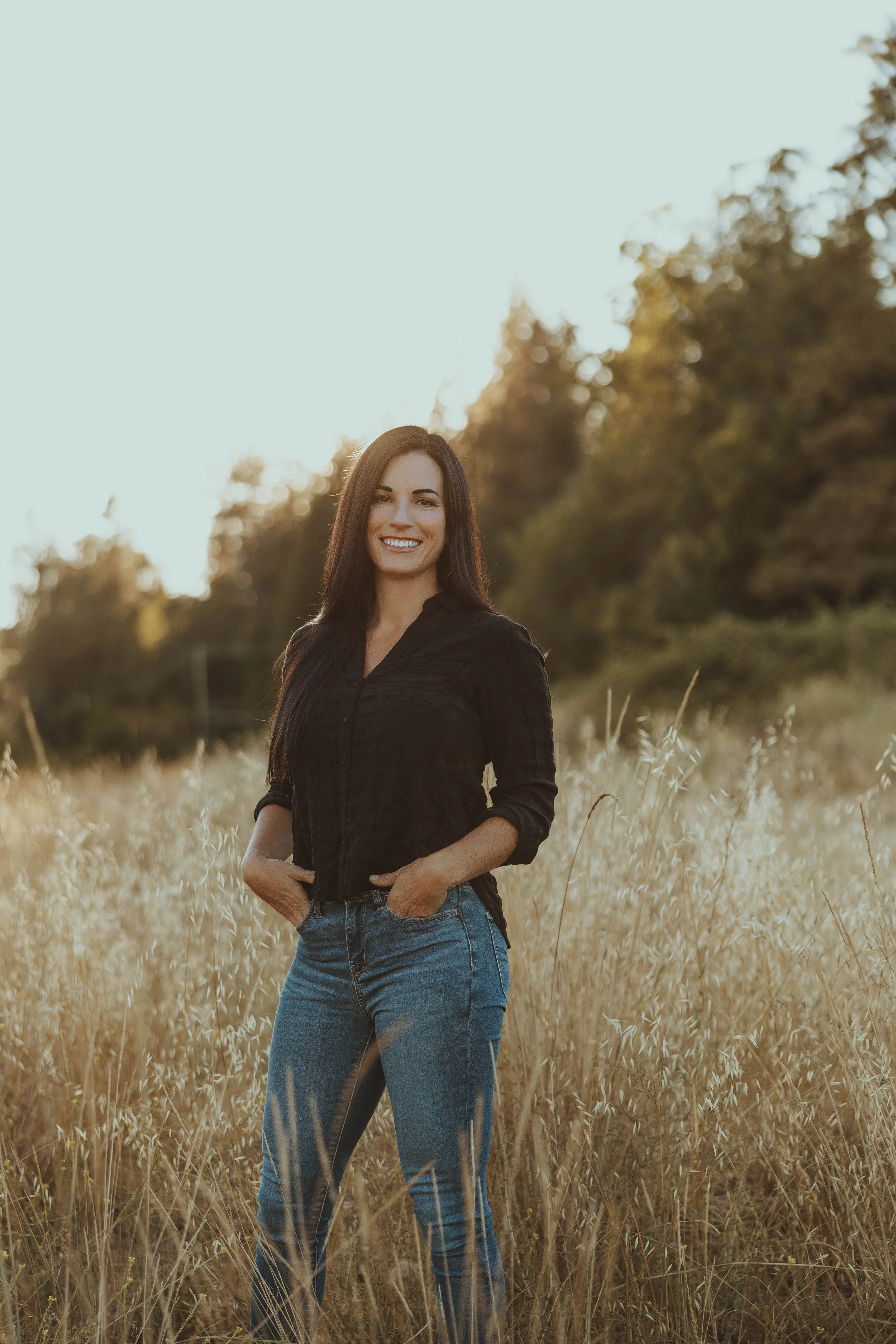 A woman standing in a field of tall grass with trees in the background, smiling at the camera during golden hour.