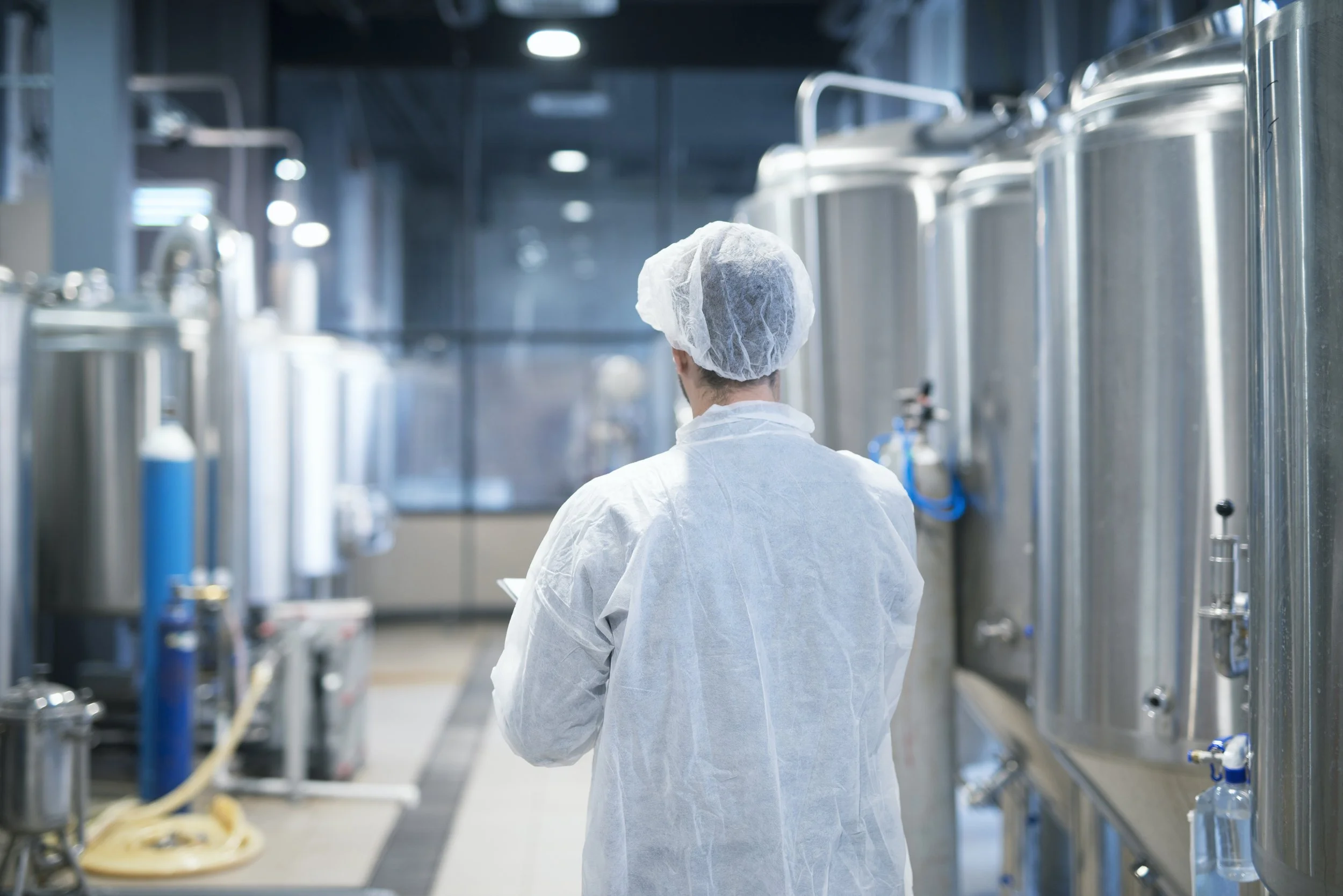 A person wearing protective clothing and a hairnet inspecting large stainless steel tanks in a clean, industrial manufacturing facility.