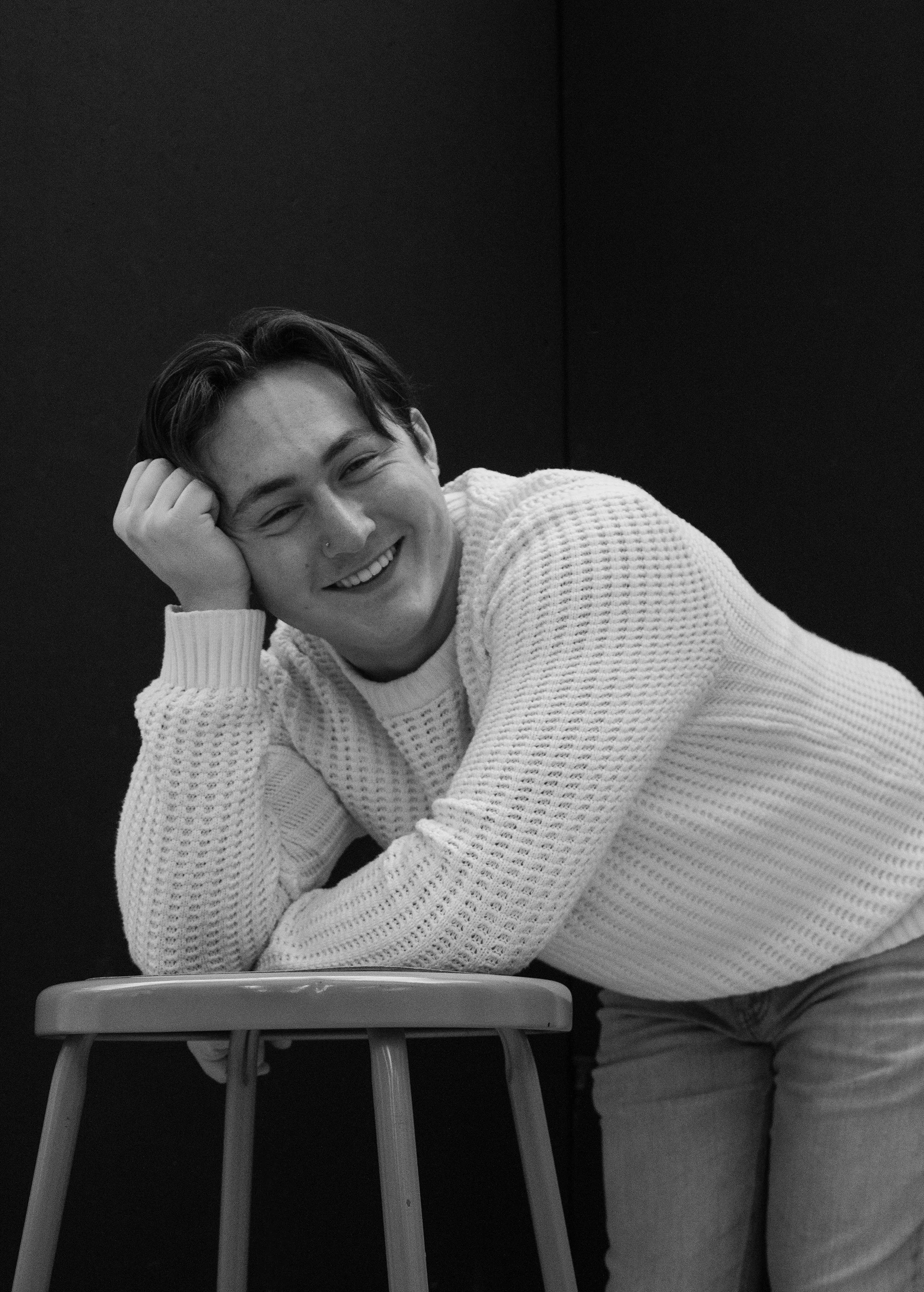 A smiling person leaning on a stool, resting their head on their hand, wearing a white knit sweater.