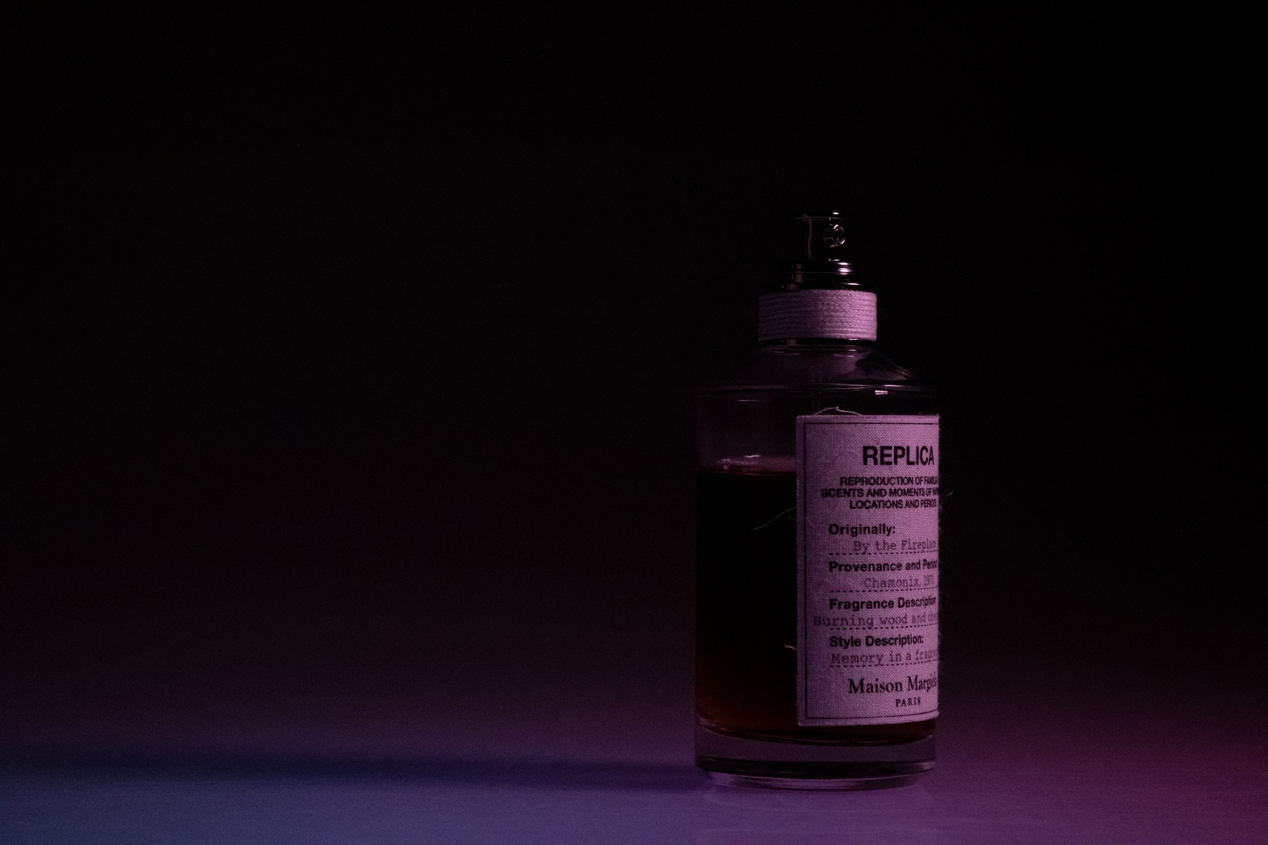 A bottle of perfume labeled 'REPLICA' with a light pink label, set against a dark background with subtle purple and pink lighting.