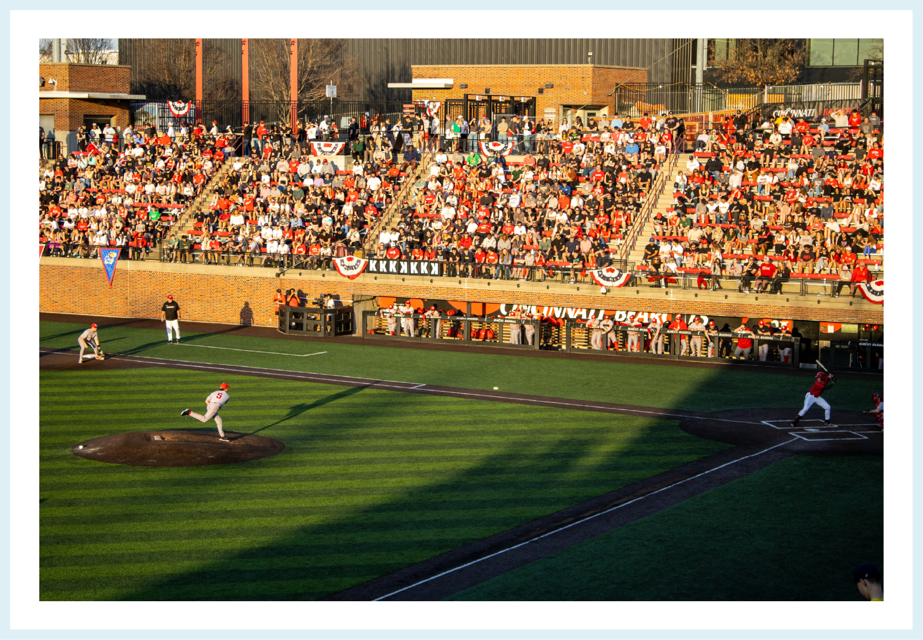 Crowd of baseball fans in stadium stands decorated with patriotic banners and flags watching a game on a sunny day. Players are on the field, with one pitcher and one batter at home plate.