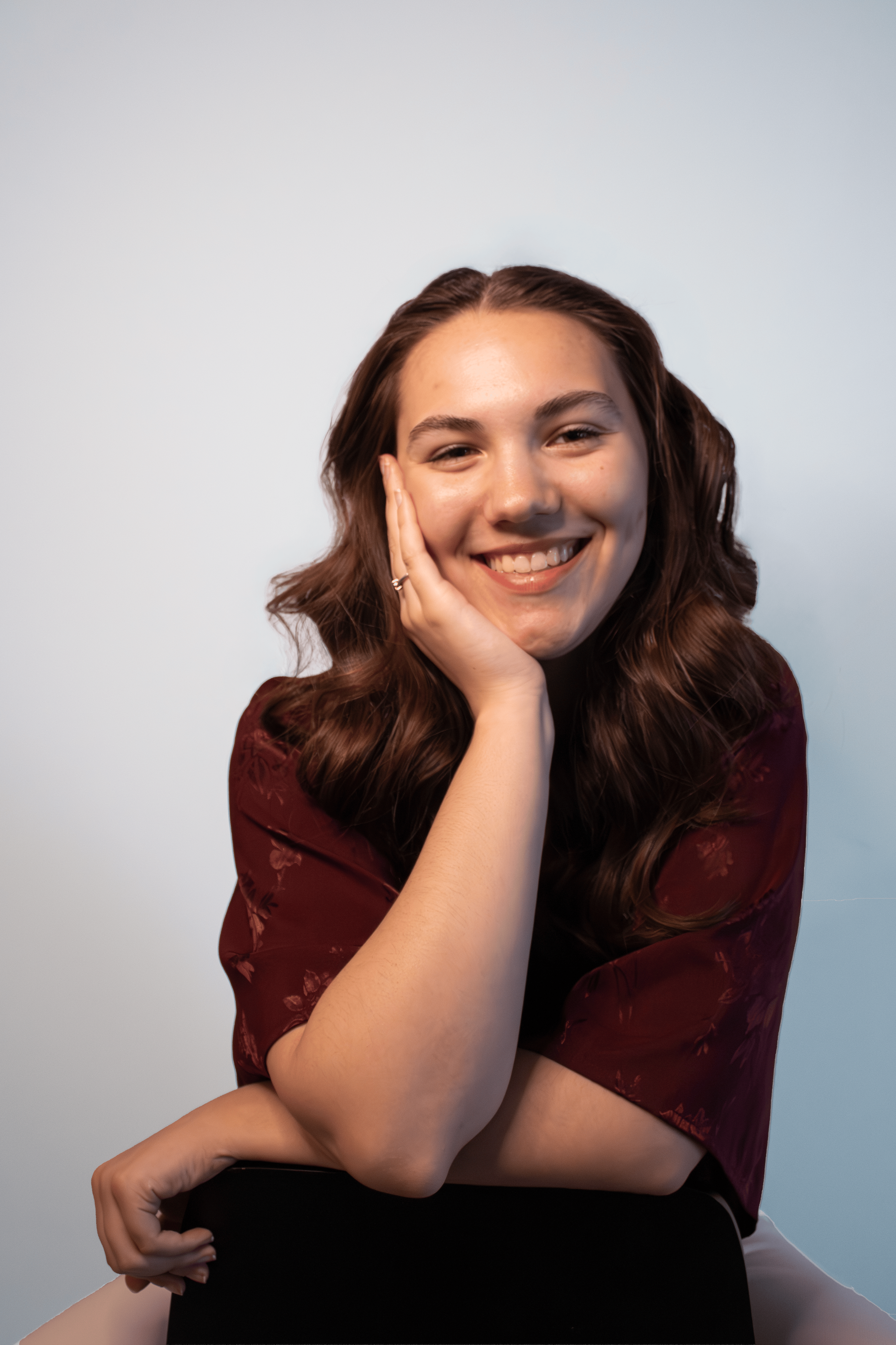 A young woman with wavy brown hair smiling and resting her head on her hand, wearing a maroon floral top against a light gray background.