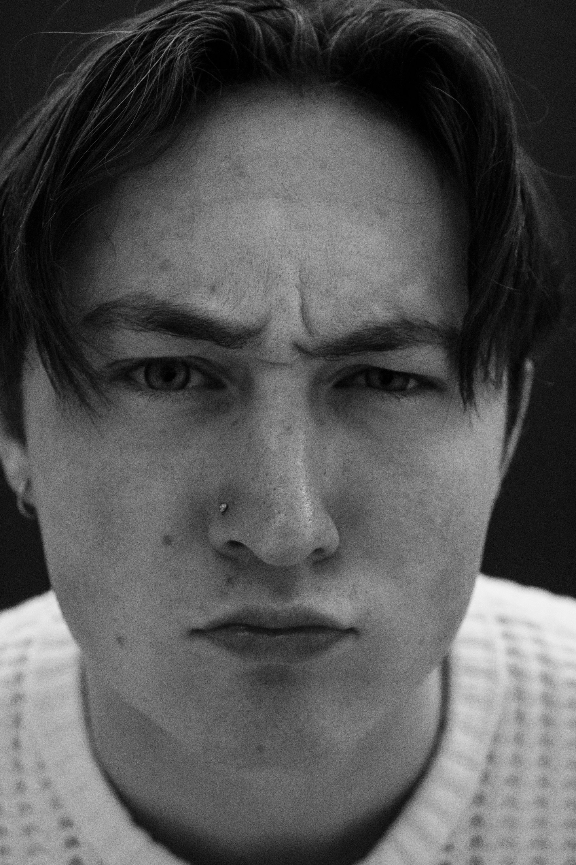 Close-up black and white portrait of a young man with dark hair, a nose piercing, and intense expression