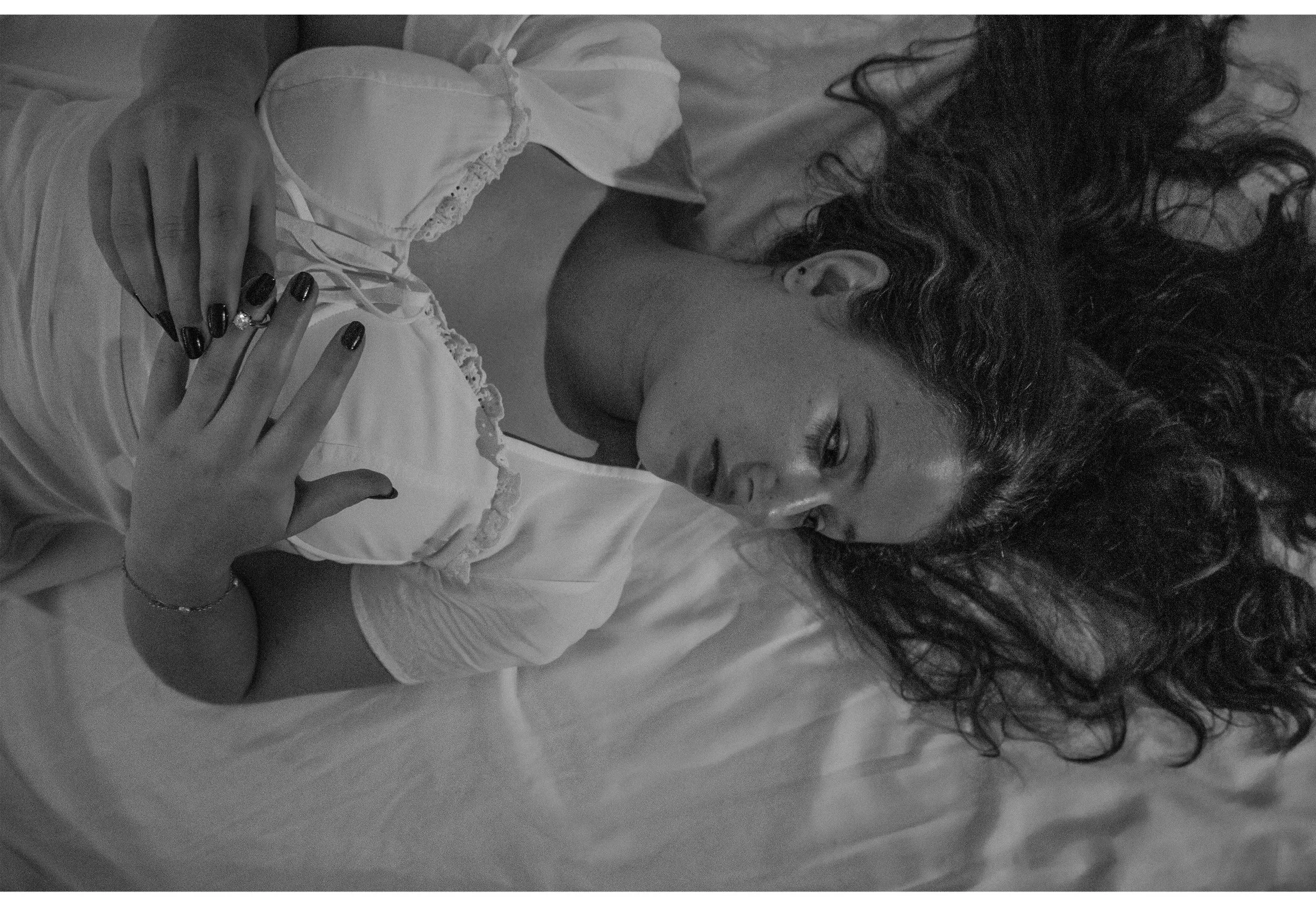 A woman with long, curly hair lying down on a bed, wearing a light-colored, satin-like garment with lace details, looking pensively to the side.