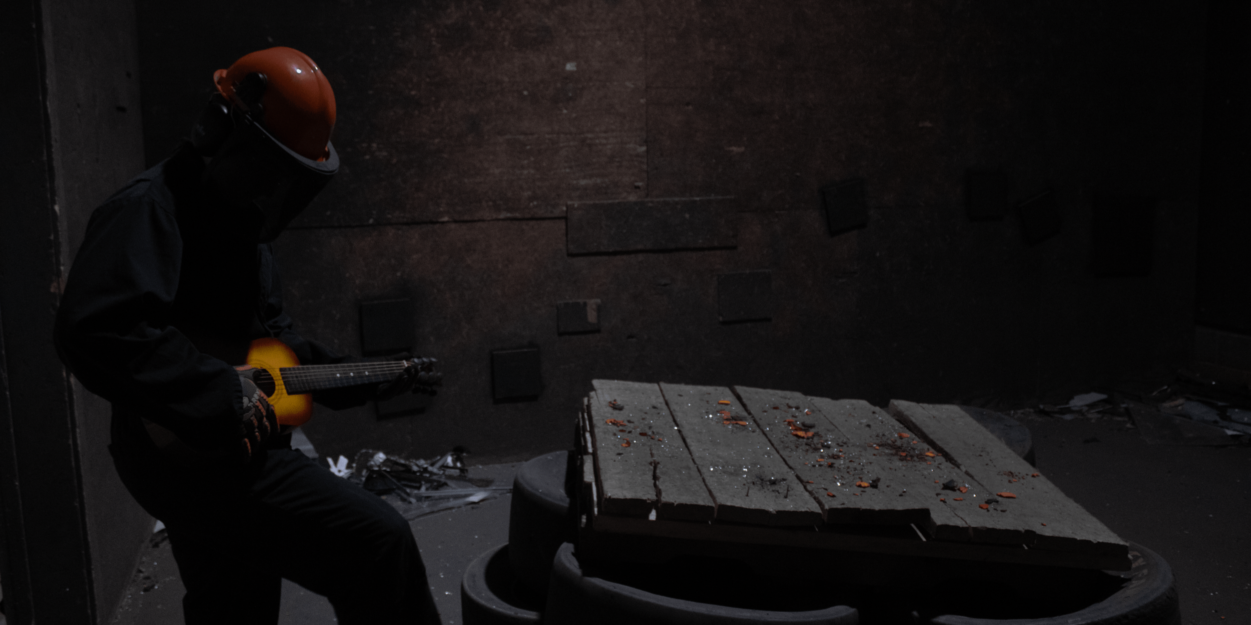 A person wearing a black jacket, dark pants, gloves, and a dark face shield and orange helmet plays an acoustic guitar in a dark room with black walls. A dirty wooden table with scattered debris is in the foreground.
