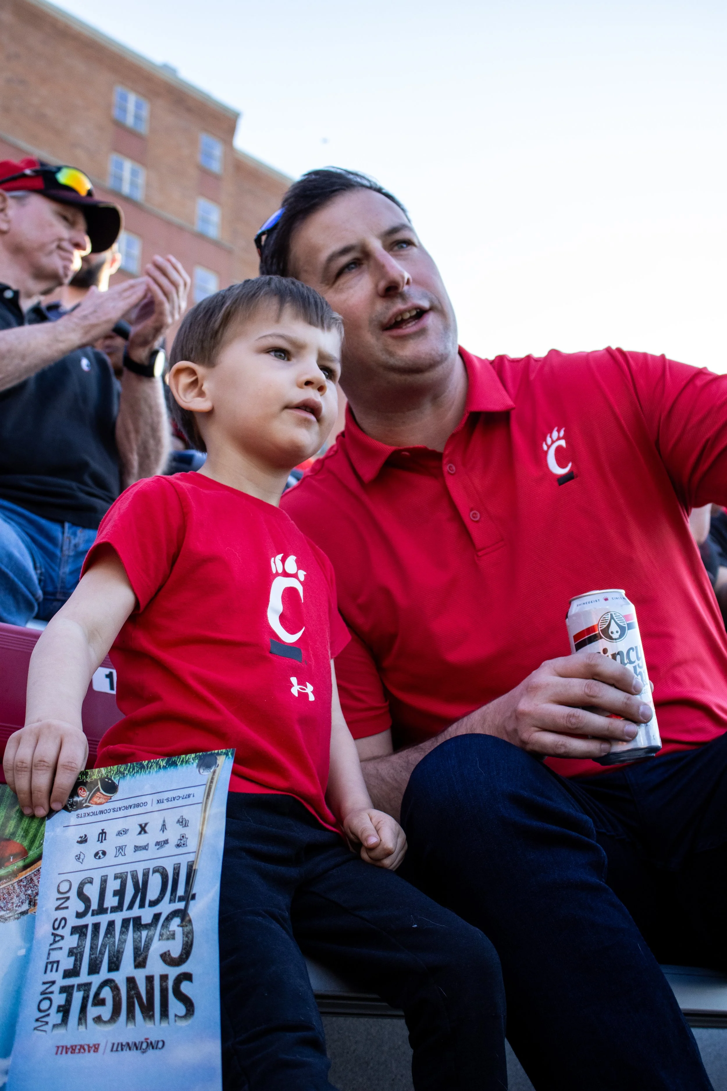 A man and a young boy sitting outdoors at a sports event, both wearing red shirts with a white 'C' logo, watching something, with the man holding a can of beer and the boy holding a printed program.
