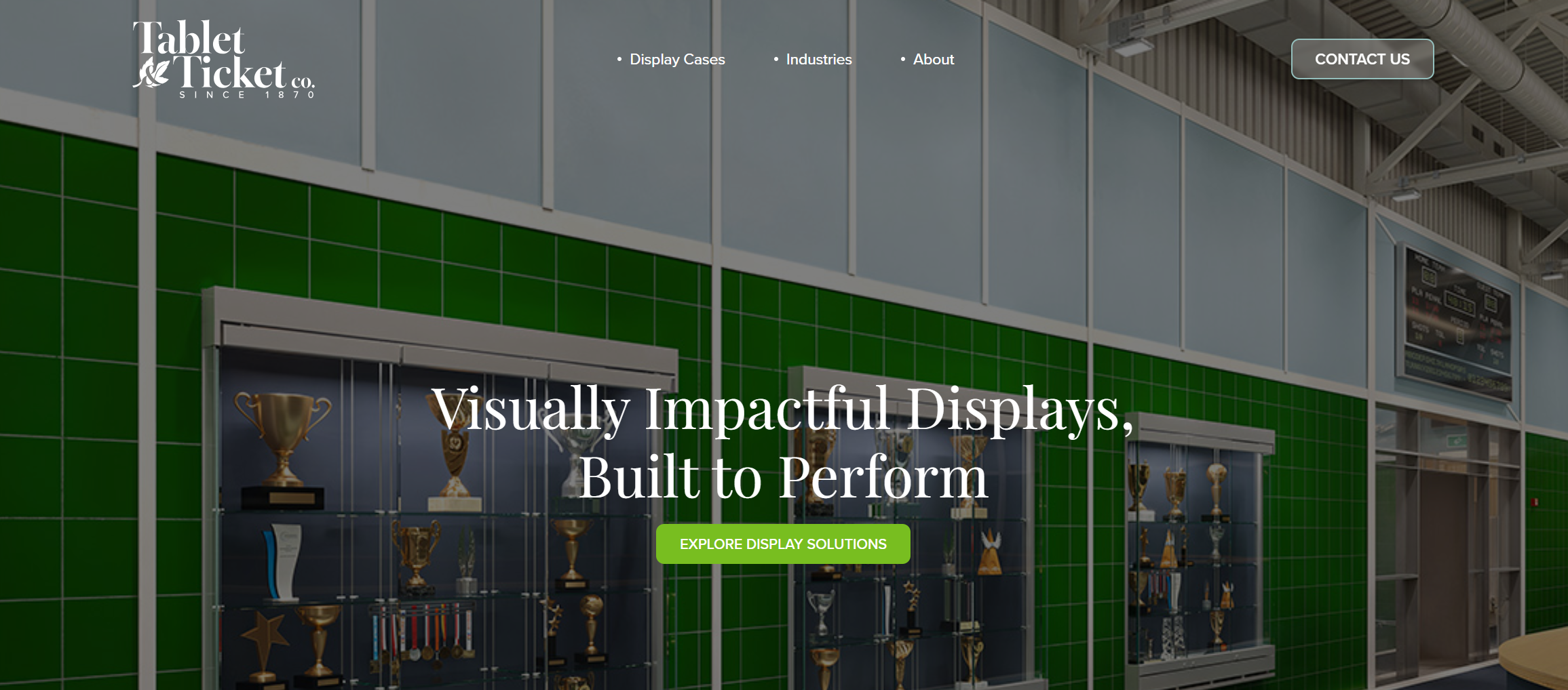 A website homepage for Tablet & Ticket Co., featuring a display of trophies in glass cases against a green tiled wall. The page has navigation links for 'Display Cases,' 'Industries,' and 'About,' with a contact button at the top right. The headline reads 'Visually Impactful Displays, Built to Perform,' with a green button labeled 'Explore Display Solutions' below.