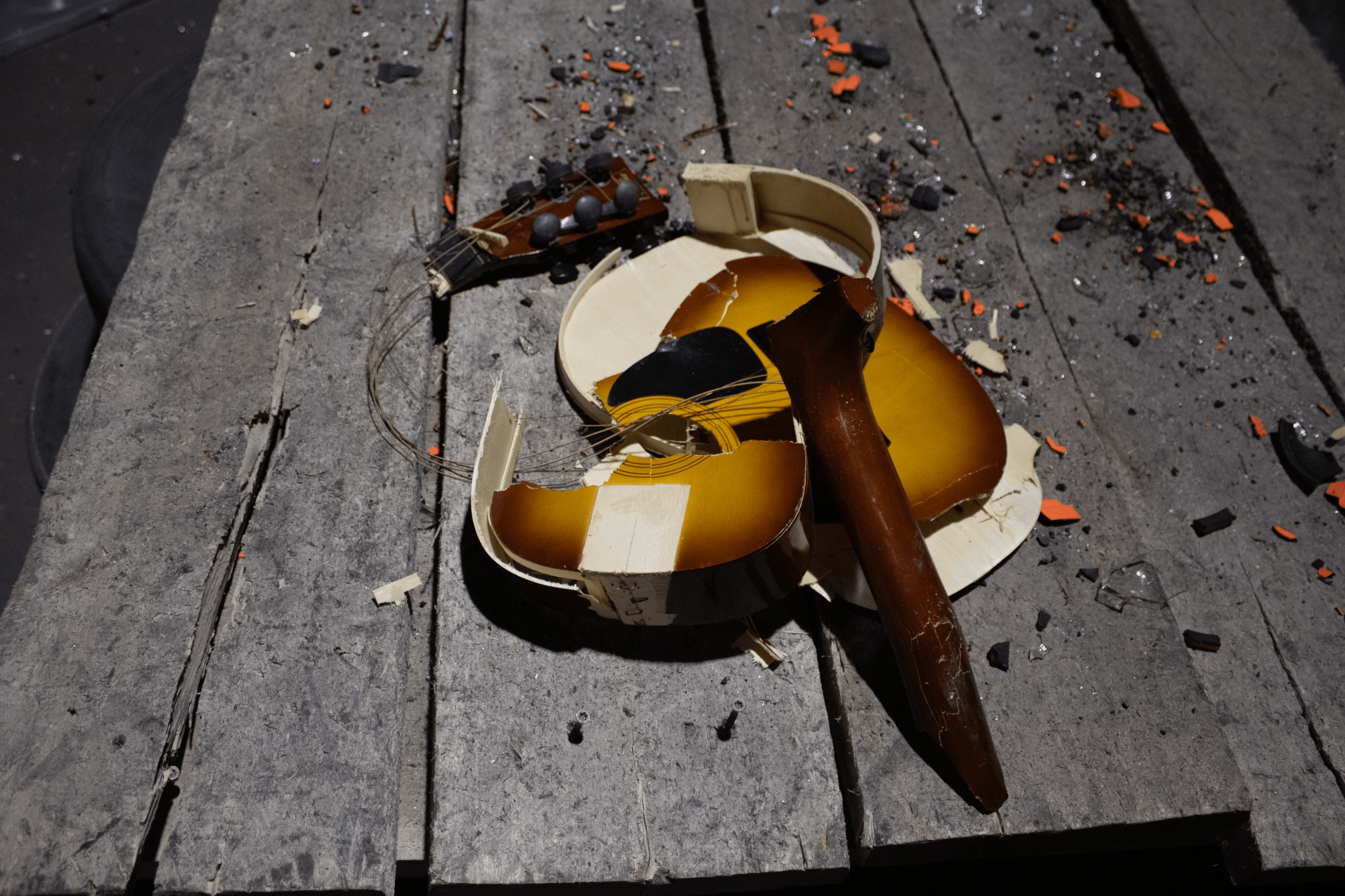 Broken guitar lying on a wooden surface with scattered debris and shattered pieces around.