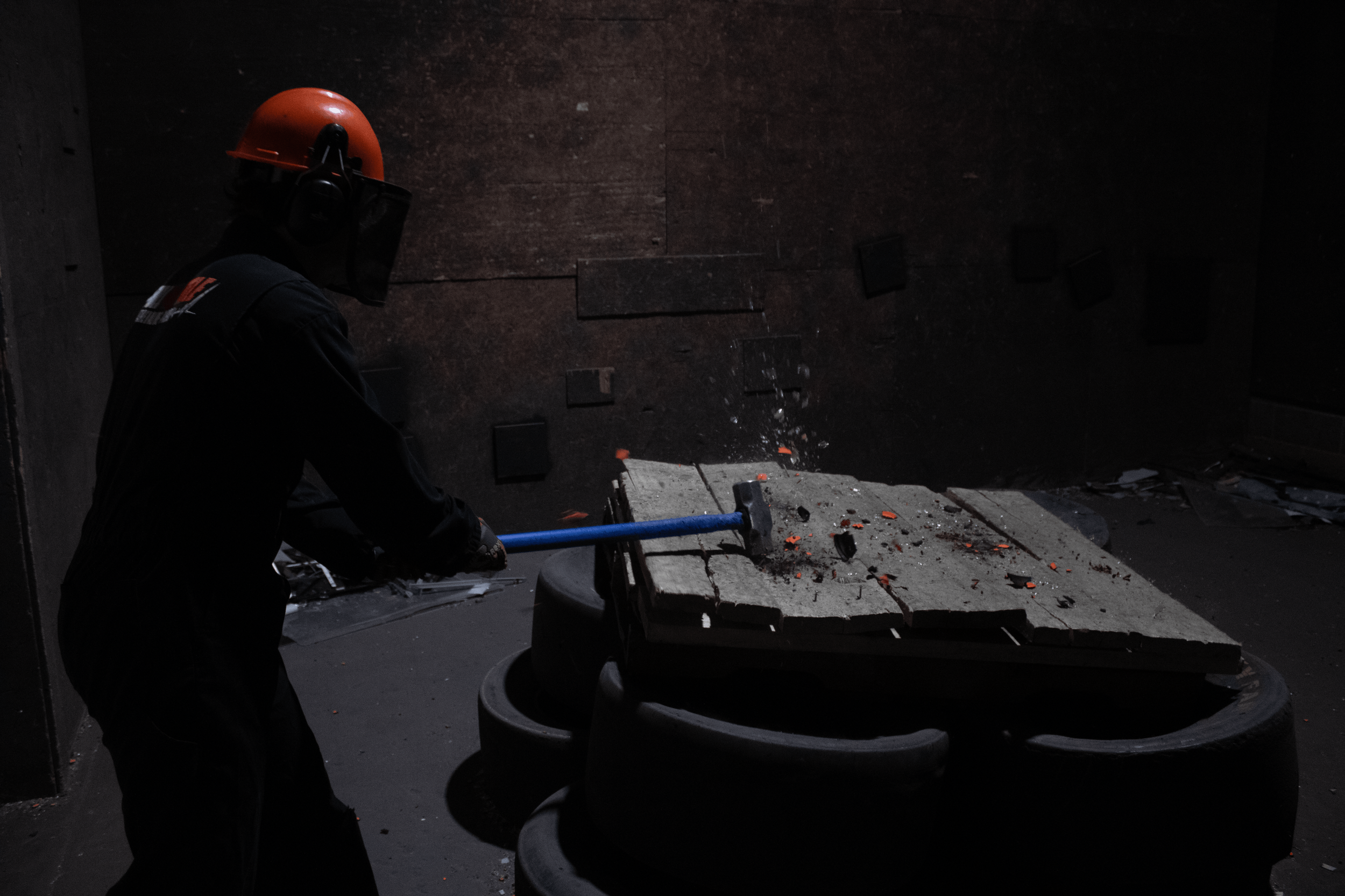 A person wearing safety gear, including an orange helmet and black clothing, is smashing a concrete slab with a sledgehammer in a dark industrial setting.